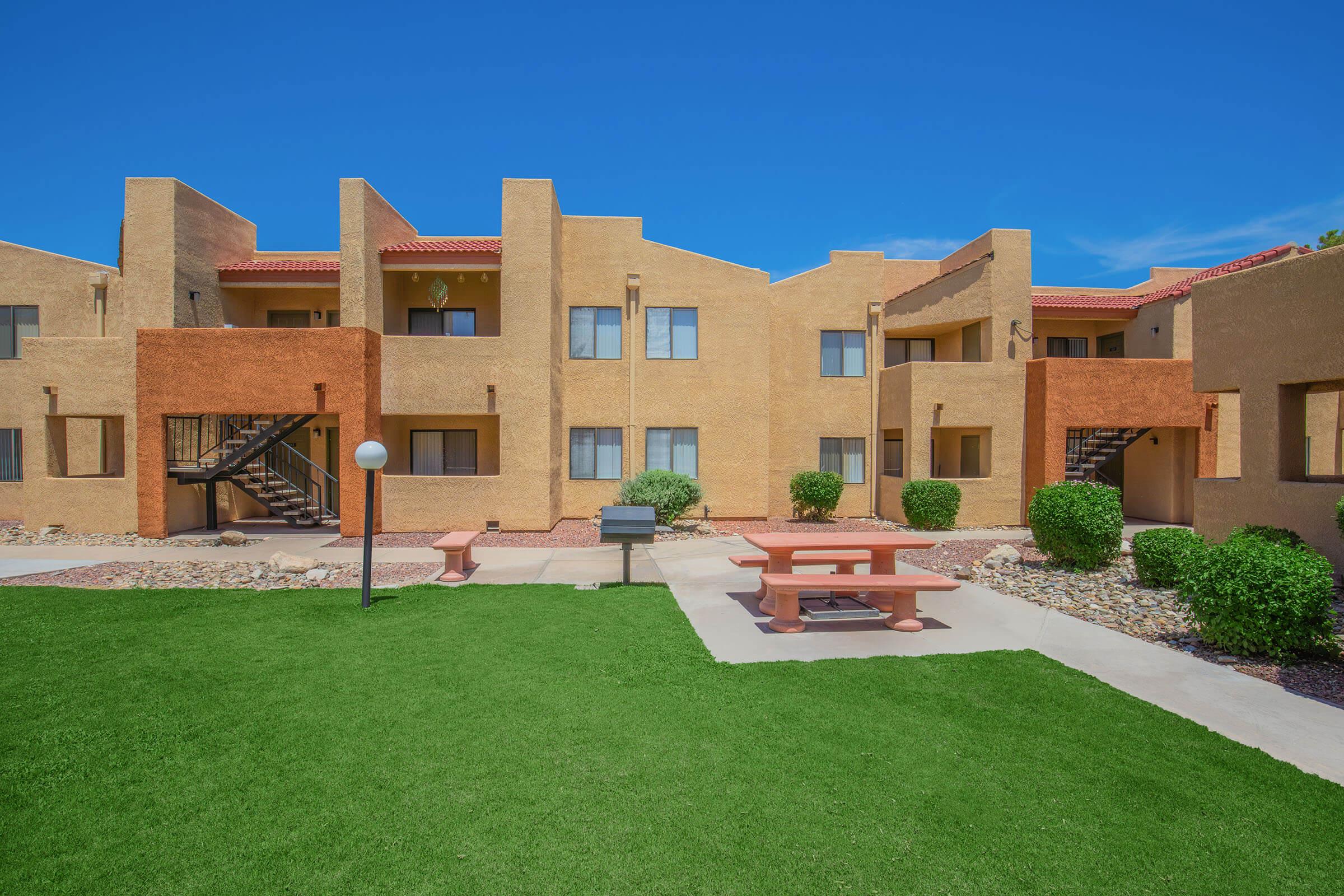 A landscaped apartment complex featuring several two-story buildings with a desert color palette. The area includes a grassy lawn, a picnic table, and light fixtures, set against a clear blue sky. The architecture showcases modern design elements with stairs leading to upper levels.