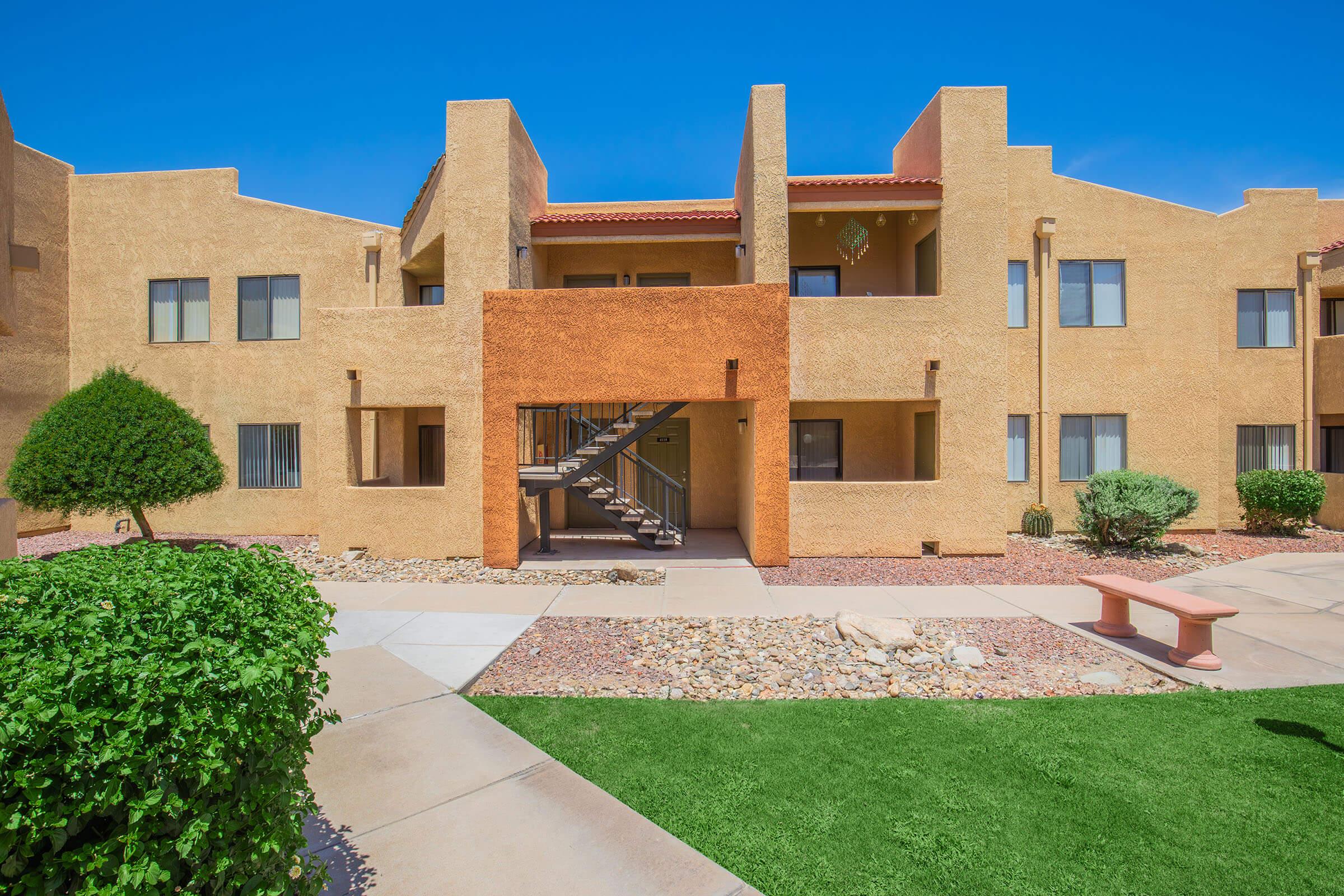A view of a multi-unit apartment building featuring a beige stucco exterior, a central staircase, and well-maintained landscaping with green shrubs and rocks. The blue sky adds a bright backdrop to the scene, creating a pleasant and inviting atmosphere.