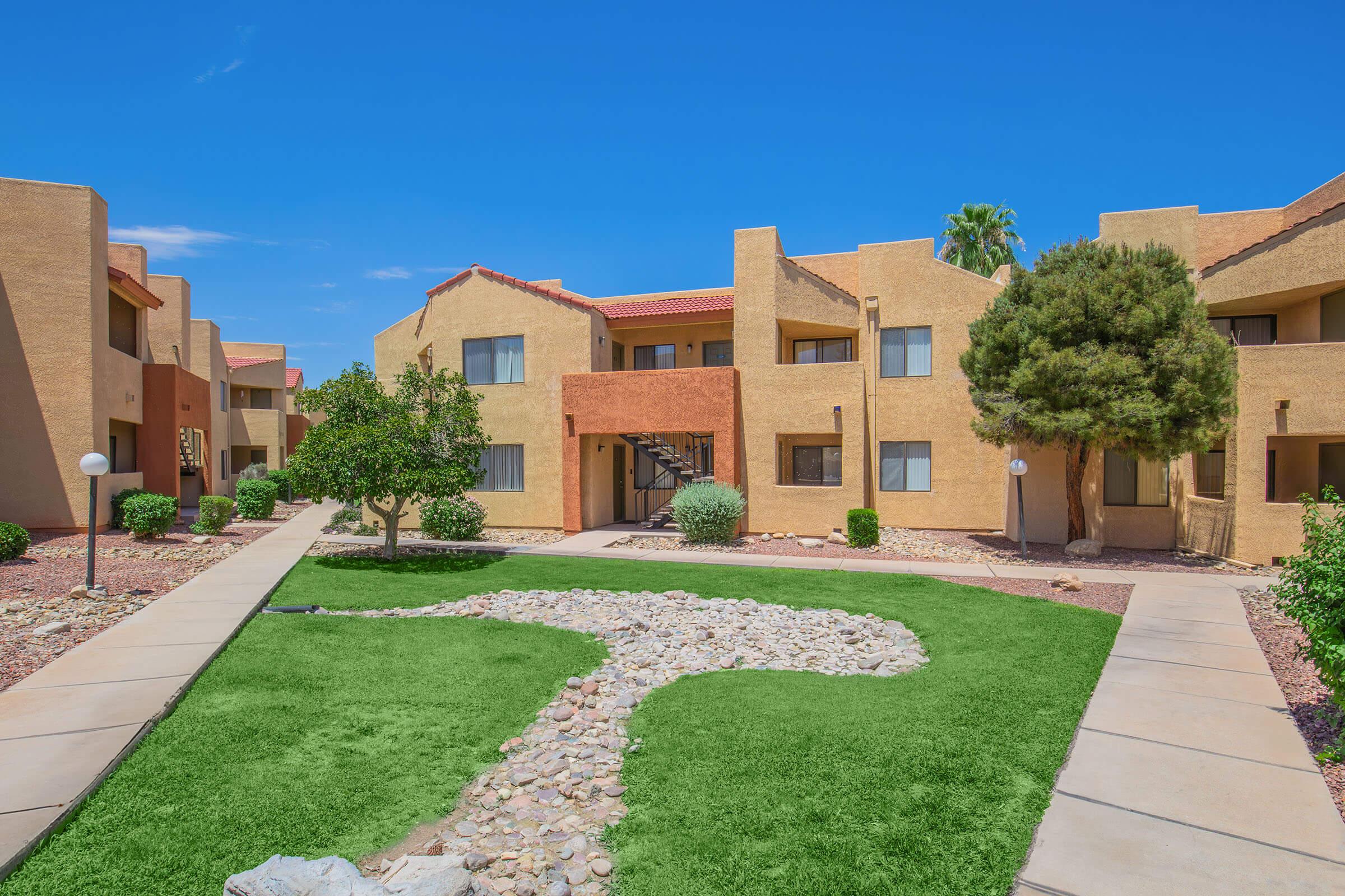 A courtyard view of an apartment complex featuring beige buildings with red tile roofs. The landscape includes green grass, a winding stone pathway, small trees, and shrubs against a clear blue sky.