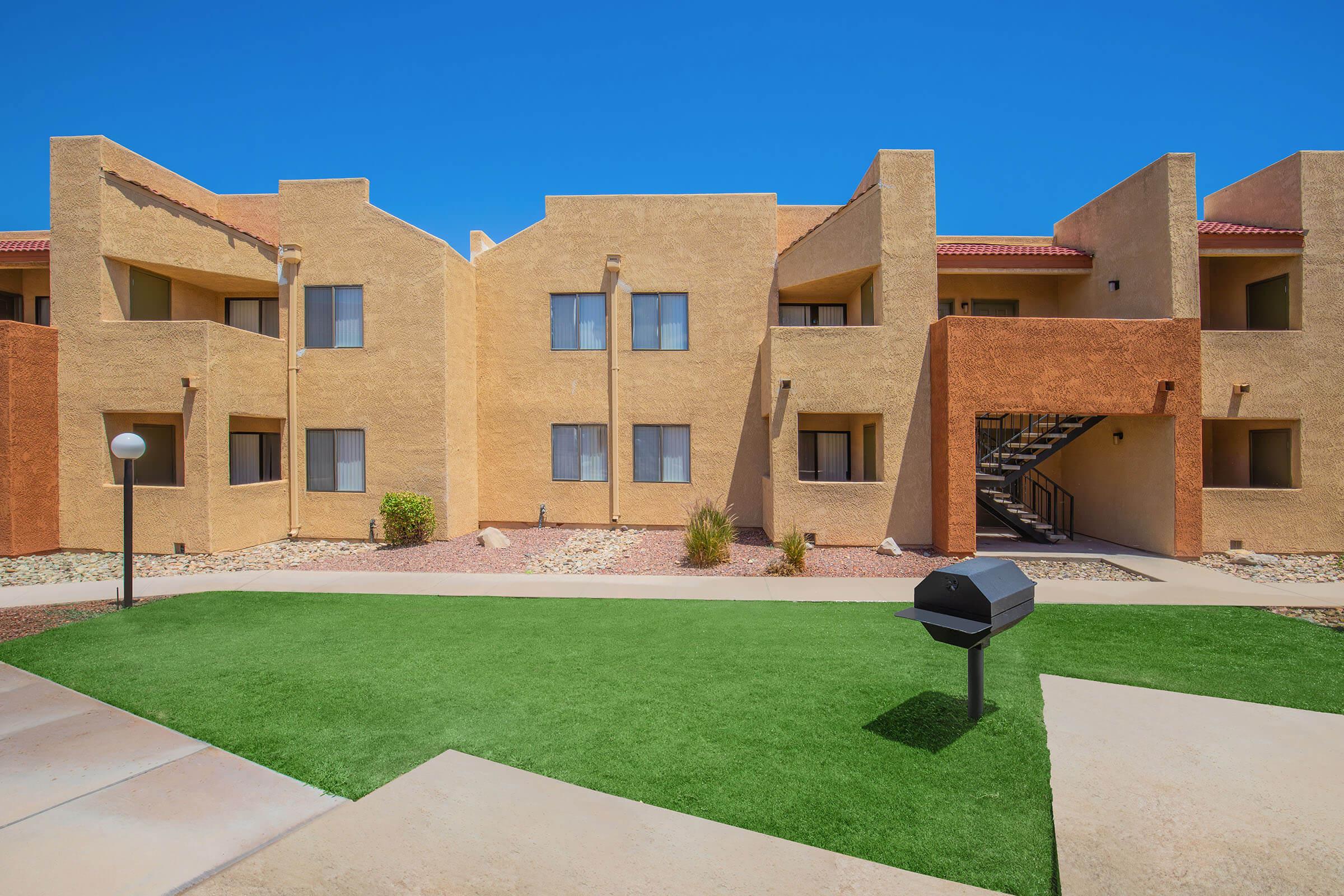 Modern apartment complex with a landscaped lawn and a black grill in the foreground. The building features a combination of tan and reddish-brown exteriors, with multiple windows and a staircase visible. Clear blue sky enhances the bright and inviting atmosphere.