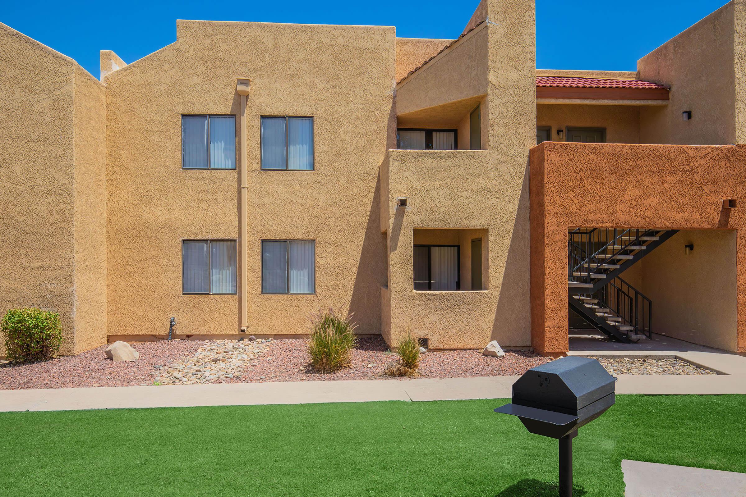 Two-story apartment complex featuring a mix of tan and orange stucco exteriors. The building has several windows with white blinds and a staircase leading to the second floor. A small grassy area is in front, with a black barbecue grill placed on a concrete path amidst low shrubs and decorative rock landscaping.