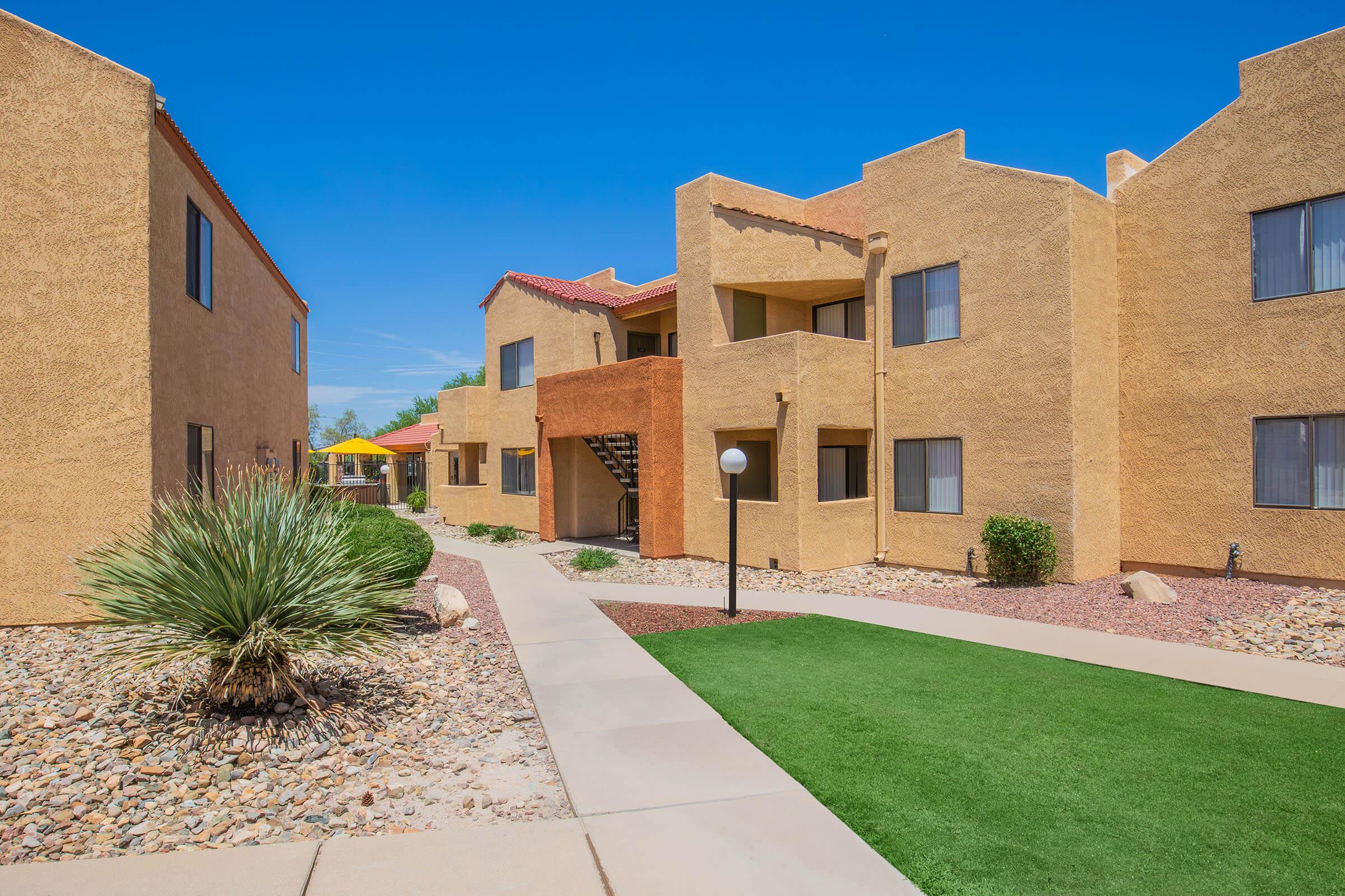 A landscaped pathway through a courtyard of two-story apartment buildings with a stucco exterior. The scene features greenery, decorative rocks, and a clear blue sky, creating a sunny and inviting atmosphere.