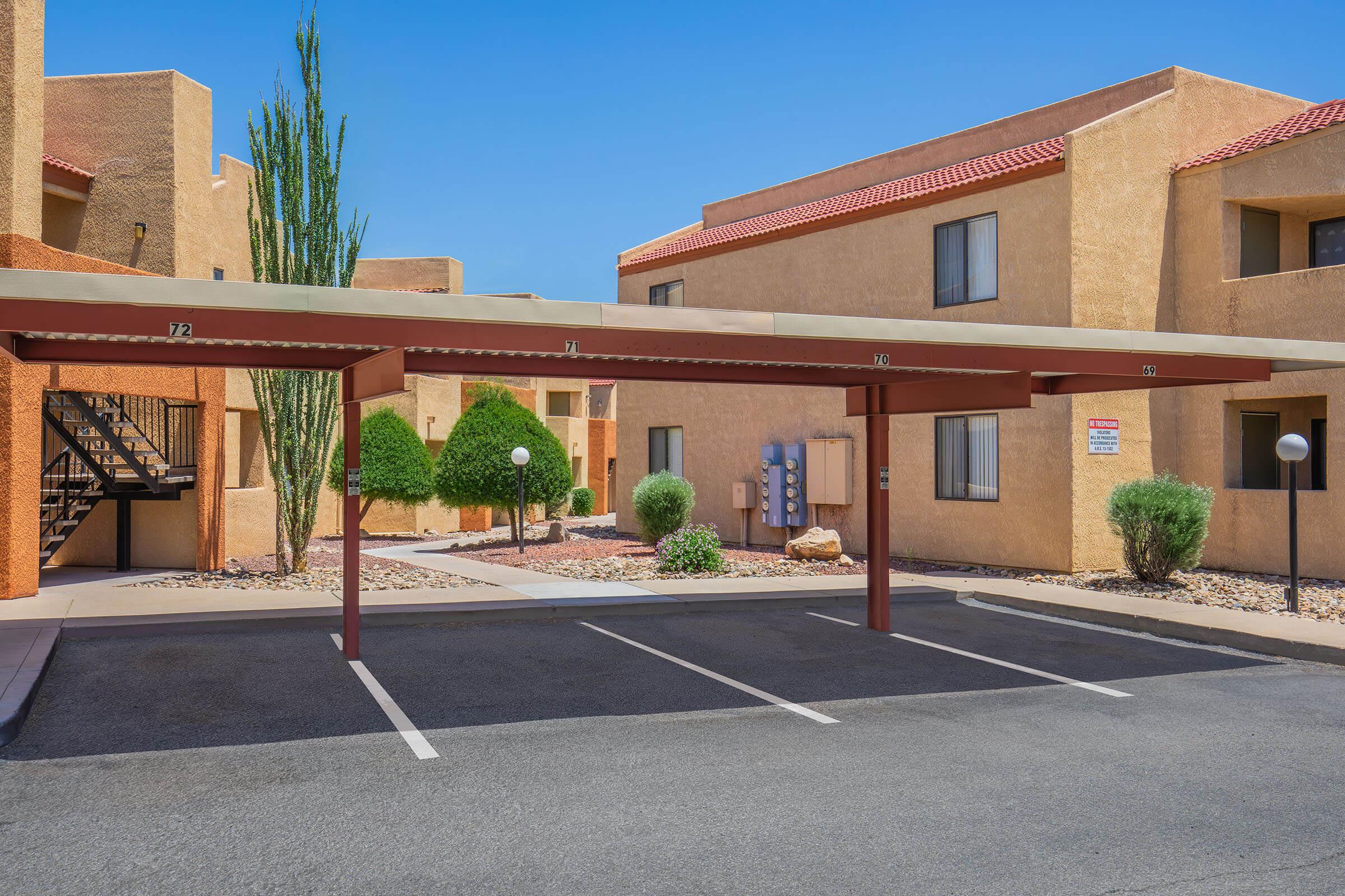 Parking area of an apartment complex featuring covered spaces, well-maintained landscaping with shrubs, and an outdoor staircase. The buildings have a warm, tan exterior with red-tiled roofs, under a clear blue sky.