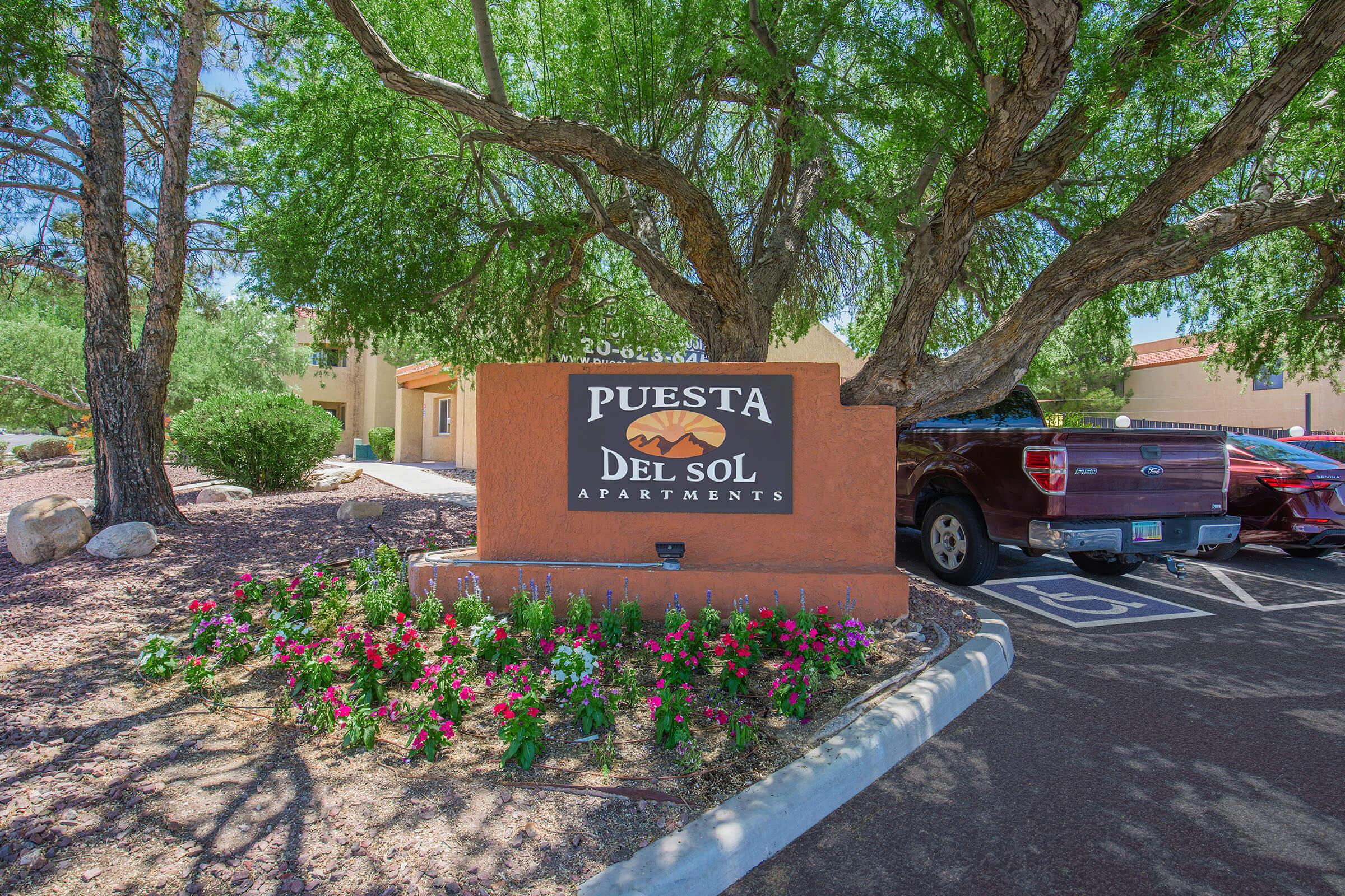 Sign for Puesta Del Sol Apartments, featuring the name prominently displayed. The sign is surrounded by colorful flowers and trees, with parked cars in the background. The scene is well-lit, showcasing a pleasant exterior in a residential area.