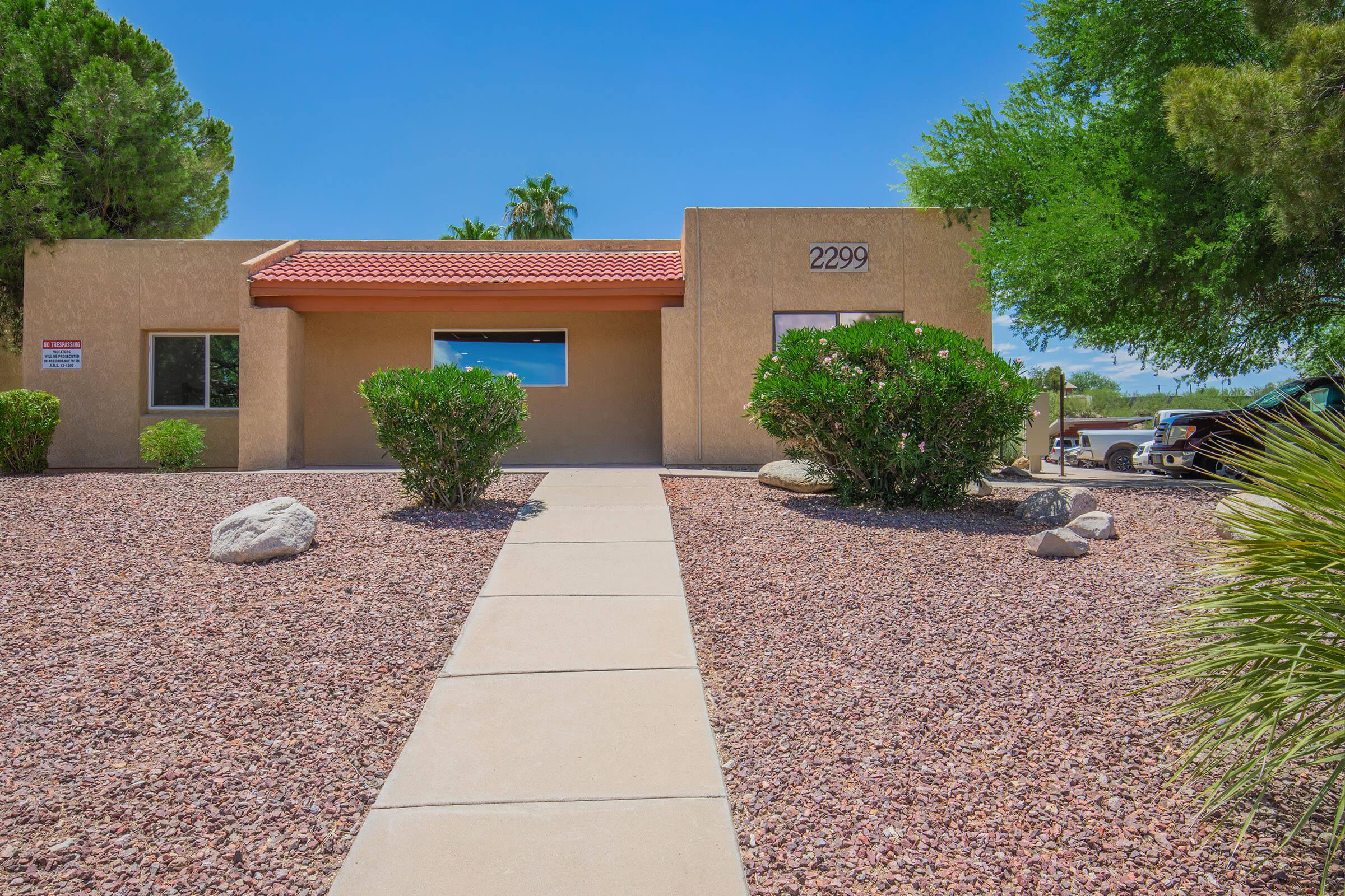 A modern single-story building with a red tile roof, surrounded by desert landscaping featuring rocks and small shrubs. The walkway leads to the entrance, marked with the number 2299. In the background, palm trees and sunny blue skies are visible.
