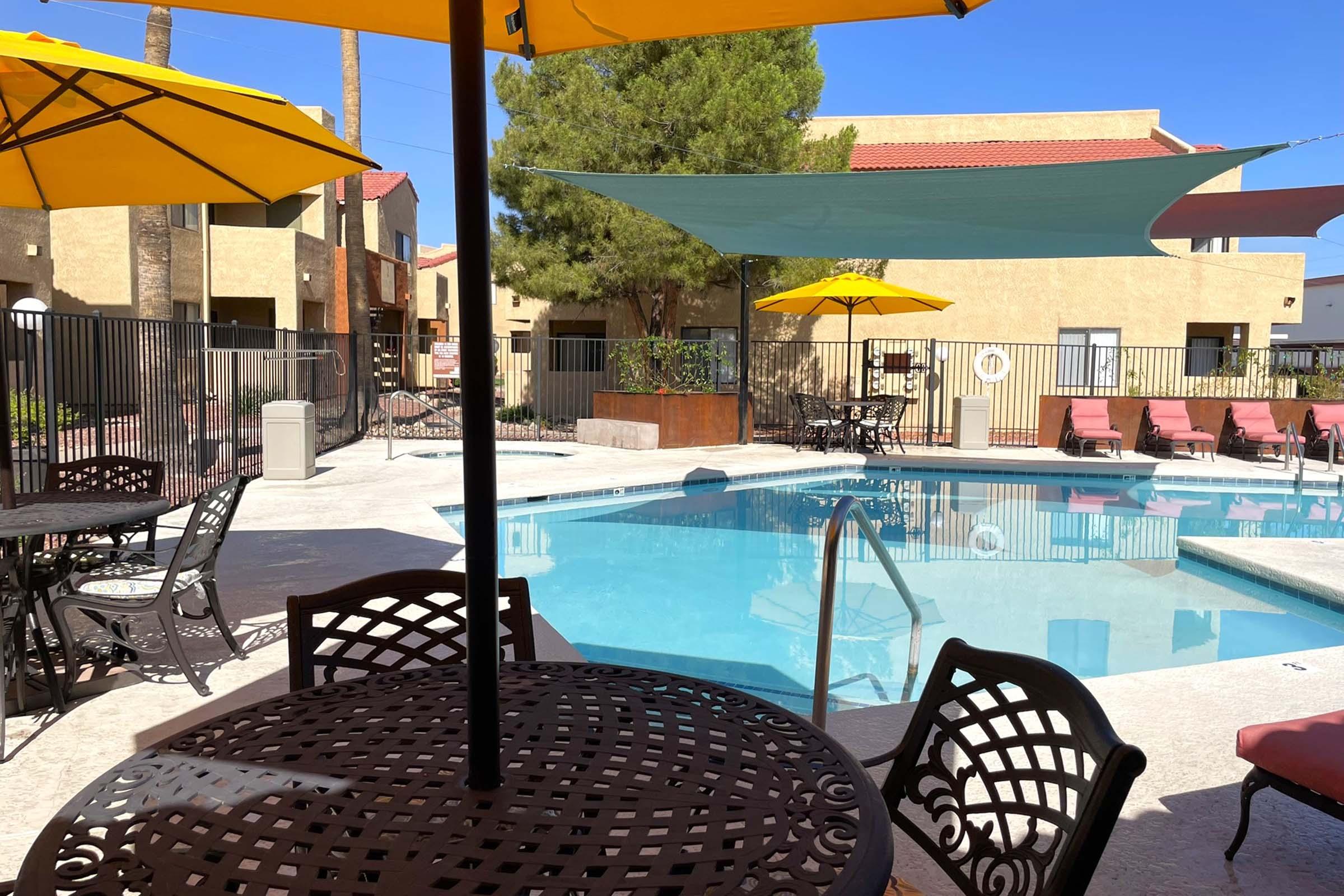 A swimming pool surrounded by lounge chairs and patio tables under yellow umbrellas. In the background, there are buildings with a desert landscape and clear blue skies. The setting offers a relaxed atmosphere for leisure and relaxation.
