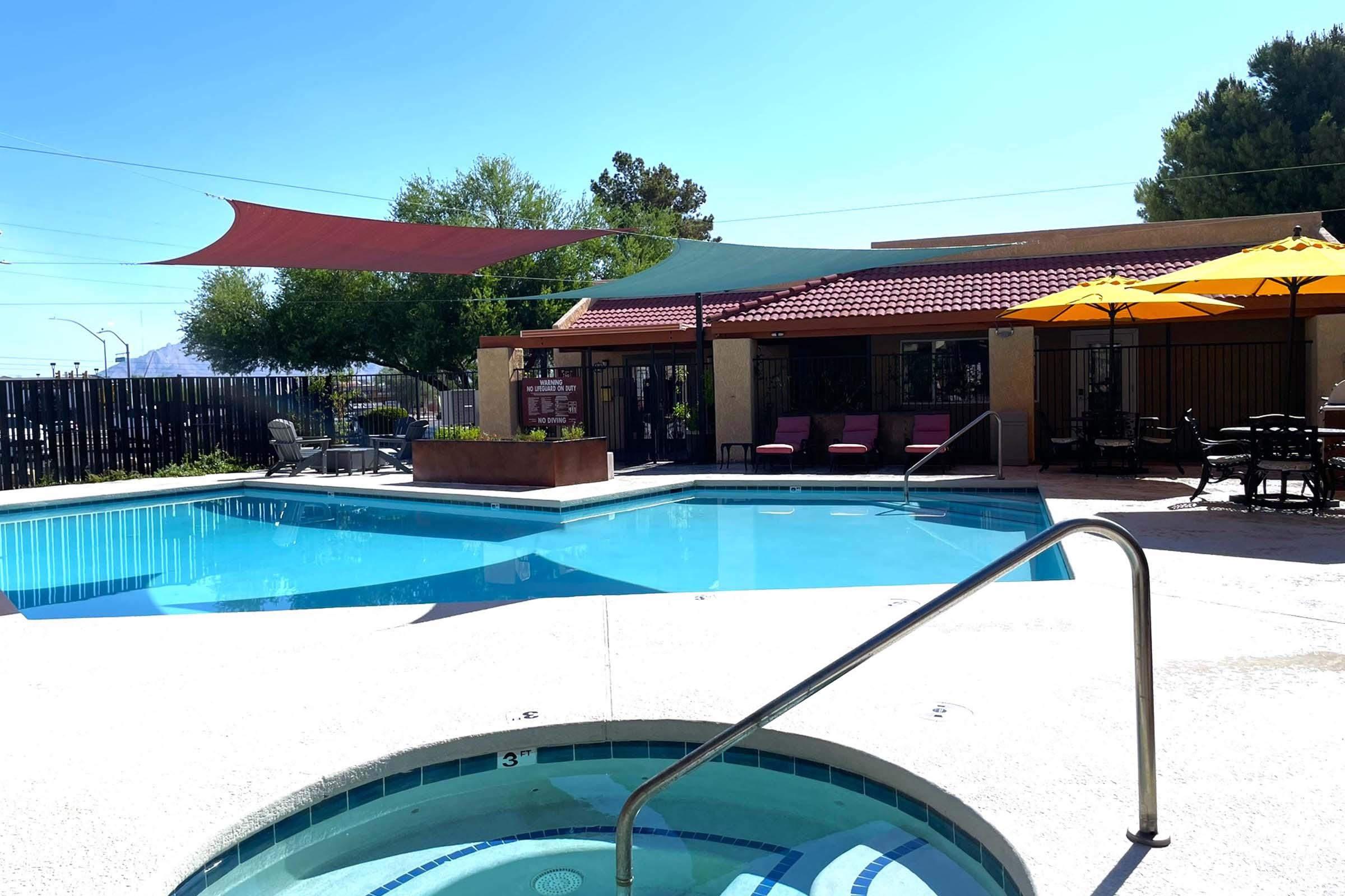 A serene pool area featuring a turquoise swimming pool with a hot tub, surrounded by sun loungers and vibrant umbrellas. Shade sails provide cover, and a well-maintained garden adds a touch of greenery. The backdrop includes a building with outdoor seating, ensuring a relaxing atmosphere. Bright blue sky enhances the inviting scene.