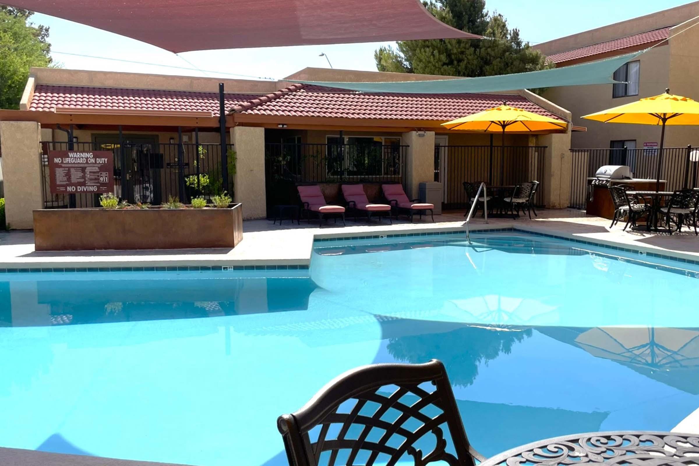 A clear blue swimming pool surrounded by lounge chairs and tables with umbrellas. The pool area is located at a building with a shaded patio and a fence. In the background, there are trees and a few outdoor furnishings, creating a cozy, relaxing atmosphere.