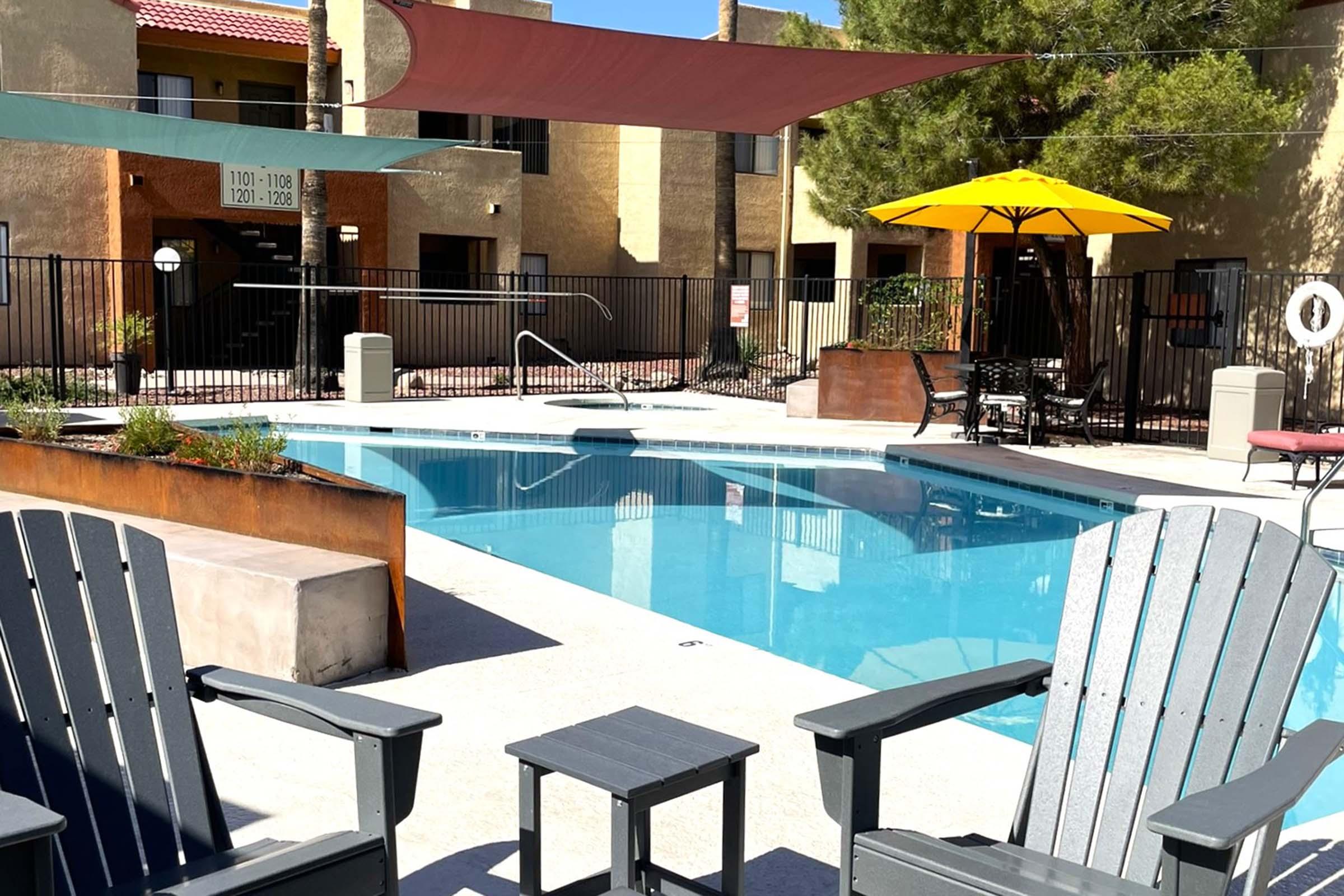 A tranquil pool area featuring a clear blue swimming pool surrounded by wooden lounge chairs and a small table. Sun umbrellas provide shade, and there are brick accents and greenery in the landscape. Nearby, there are apartment buildings in a sunny, landscaped setting.