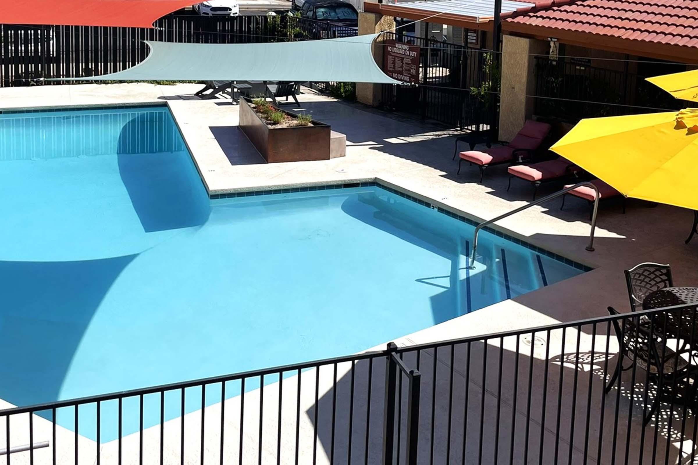 A bright outdoor pool area featuring a crystal clear swimming pool, surrounded by lounge chairs. Colorful umbrellas provide shade, and a landscaped planter is situated beside the pool. The area is enclosed by a black metal fence, suggesting a relaxing atmosphere in a sunny environment.