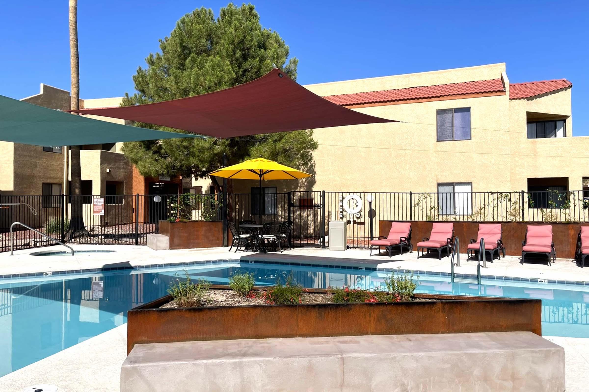 A swimming pool surrounded by lounge chairs with shaded areas created by colorful canopies. In the background, there are apartment buildings and a large yellow umbrella near the poolside. The scene is bright and sunny, with greenery and decorative plants enhancing the ambiance.