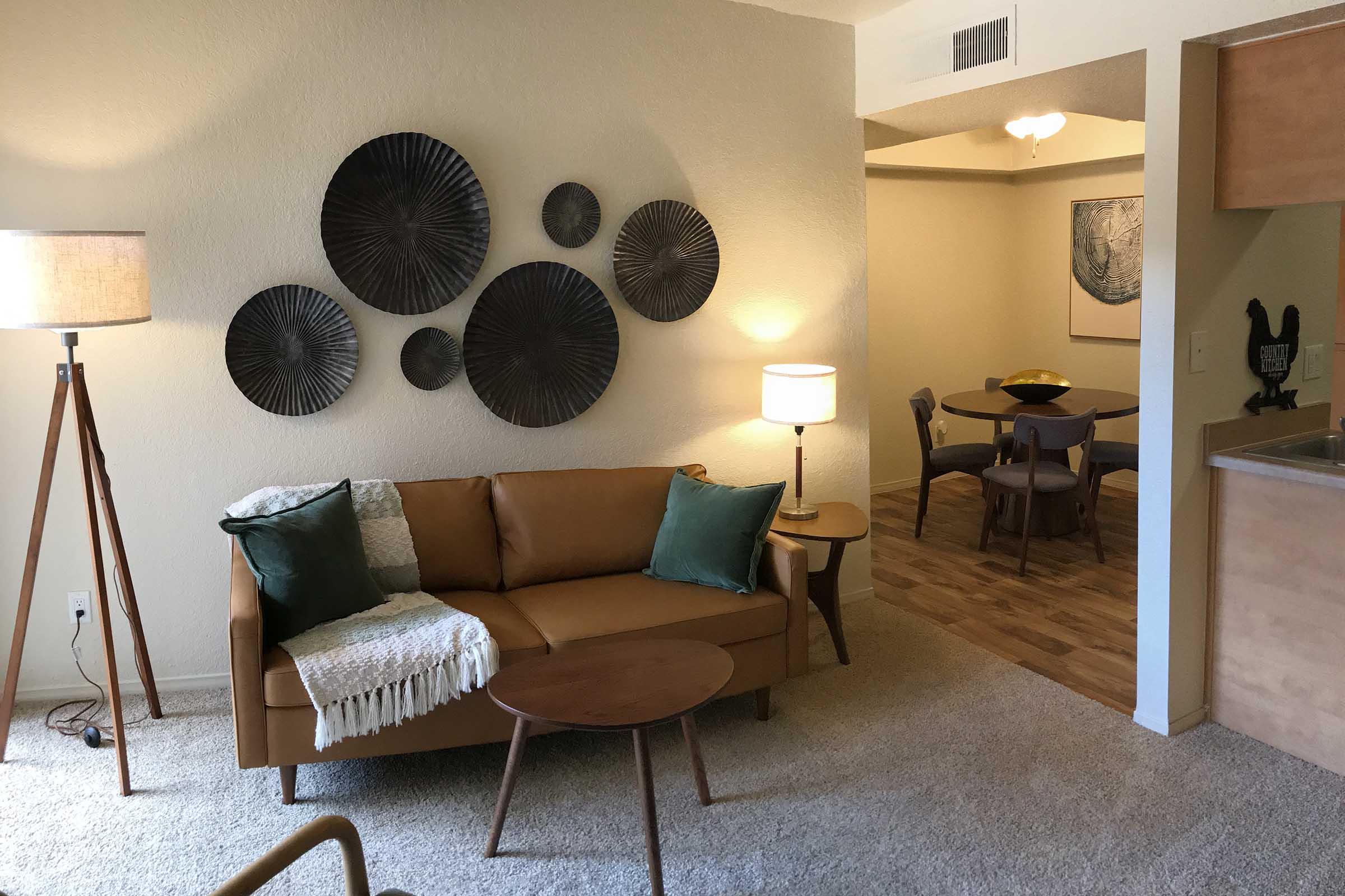 A cozy living room featuring a brown leather sofa with green accent pillows and a textured throw blanket. On the wall, there are several decorative circular wall sculptures. A standing lamp and a small round table are nearby. In the background, a dining area with a round table and chairs is visible.