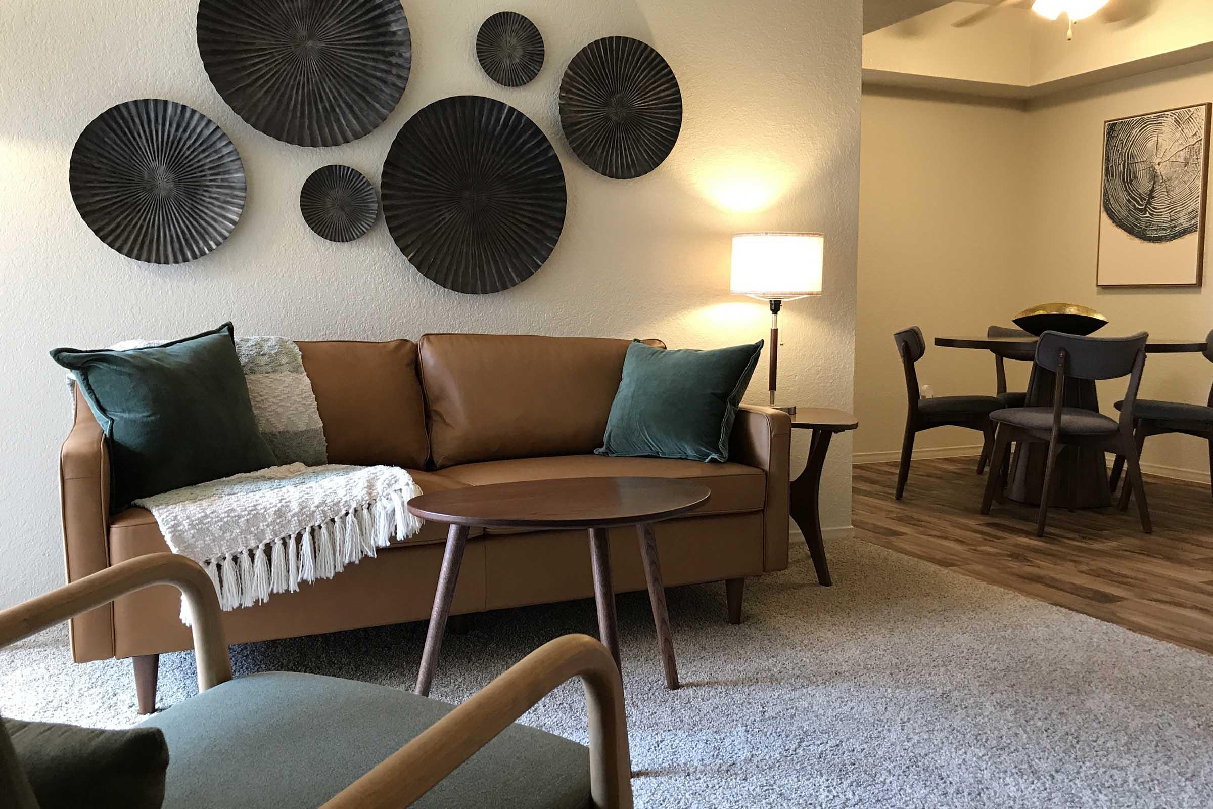 A cozy living room featuring a brown leather sofa adorned with green and white cushions. A wooden coffee table sits in front of the sofa, and a decorative wall display of circular black artworks hangs above. A floor lamp provides soft lighting, and a dining area with a round table and chairs is visible in the background.