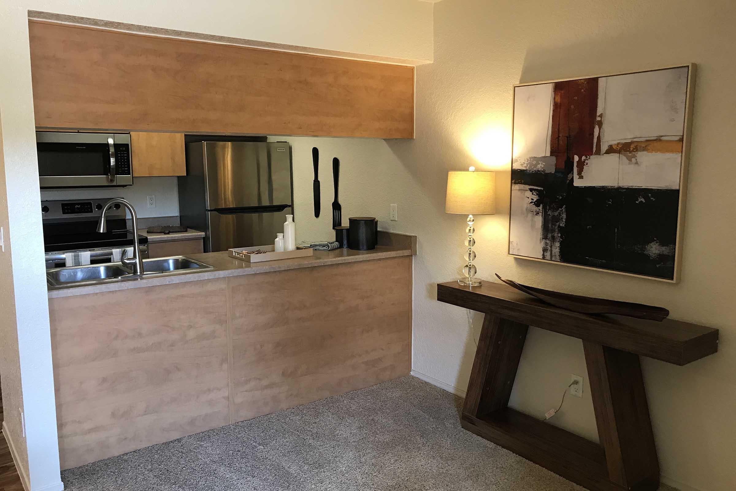 A modern kitchen space featuring a bar-style countertop with a sink, a black lamp, and an abstract painting on the wall. The kitchen includes stainless steel appliances, wooden cabinetry, and utensils hanging near the sink. The area has carpeted flooring and a warm, inviting atmosphere.