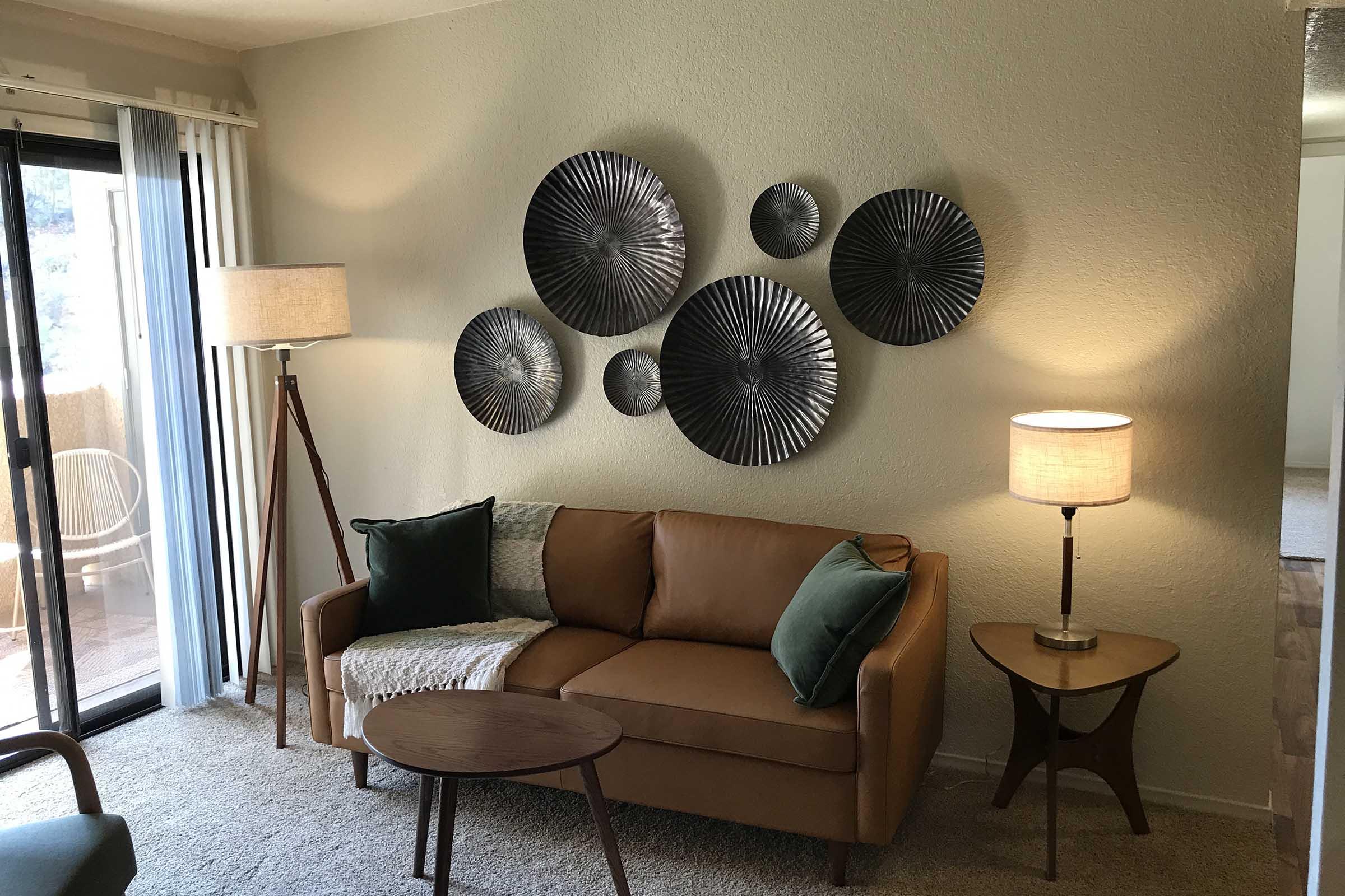 Modern living room featuring a brown couch with green and white throw pillows, a small round wooden coffee table, and a floor lamp. The wall is decorated with metal wall art in circular designs. A side table with a lamp is also present, and there are sliding glass doors leading to an outdoor space.