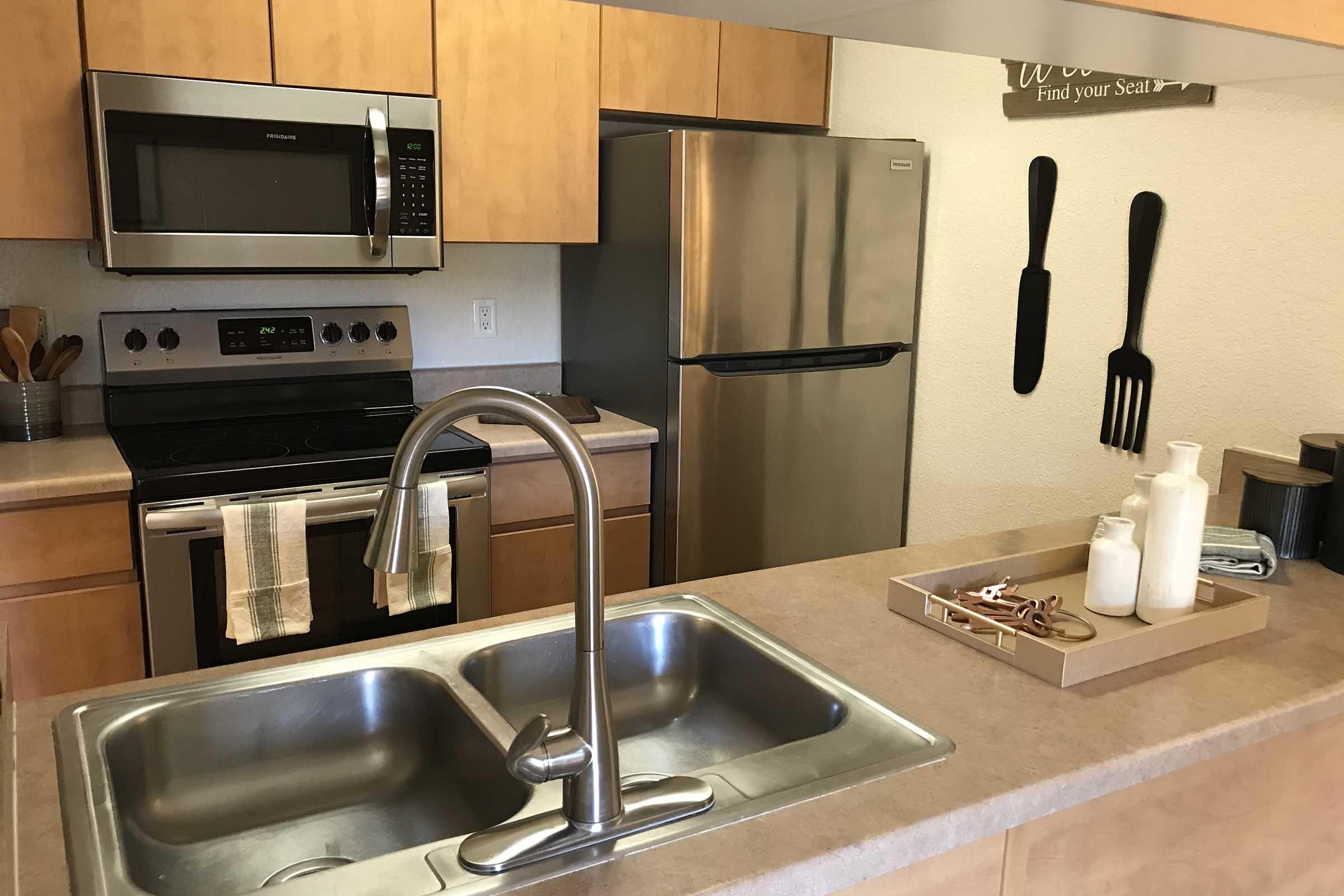 A modern kitchen featuring wooden cabinets, a stainless steel refrigerator, microwave, and stove. A sink with a sleek faucet sits in the foreground, with a tray holding decorative items. Wall decor includes large kitchen utensils and a sign that reads, "Find your seat."