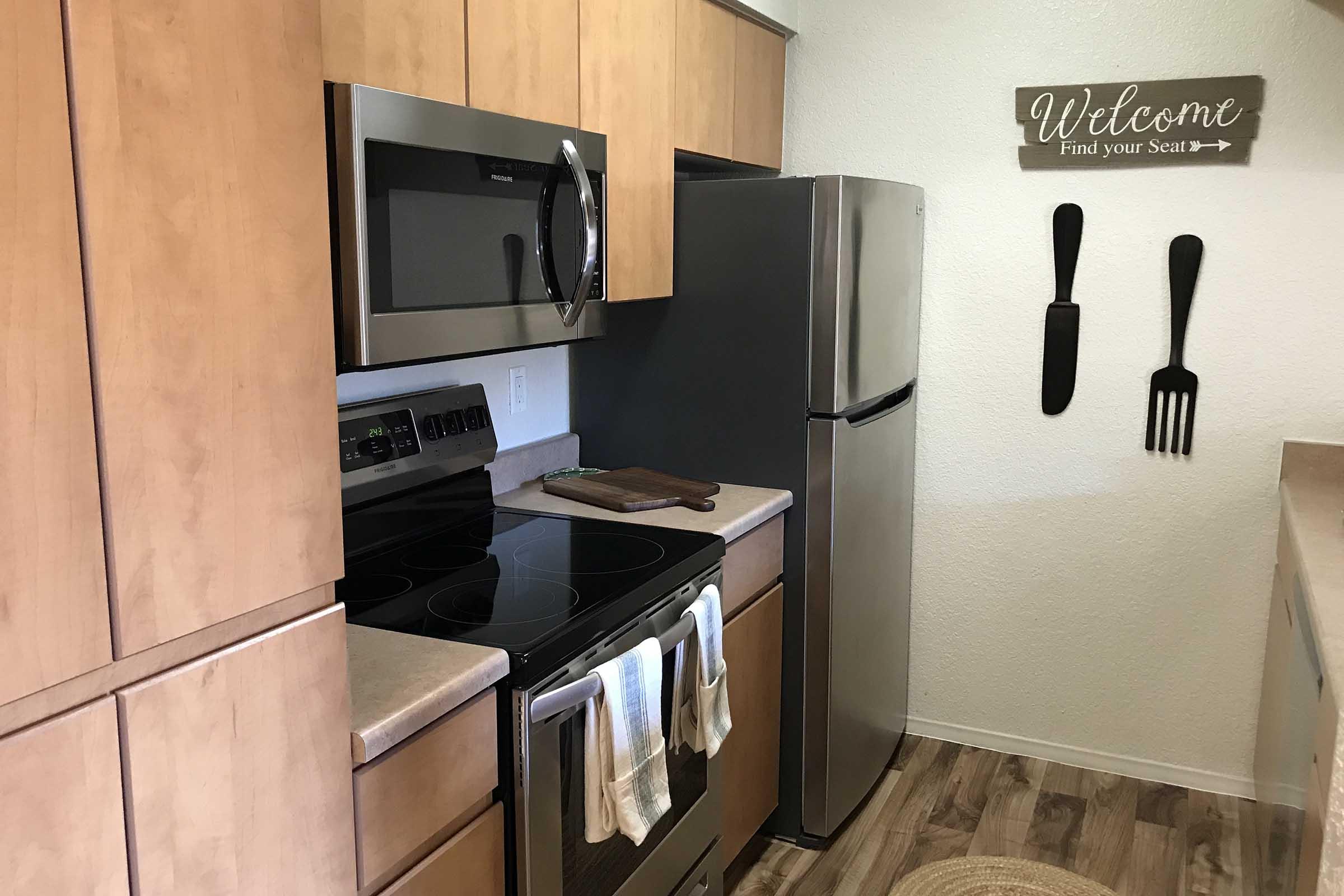 A modern kitchen featuring wooden cabinets, stainless steel appliances including a microwave and refrigerator, and a black stove. There’s a cutting board on the countertop and a decorative sign that reads "Welcome, Find your Seat" alongside large utensils hanging on the wall.