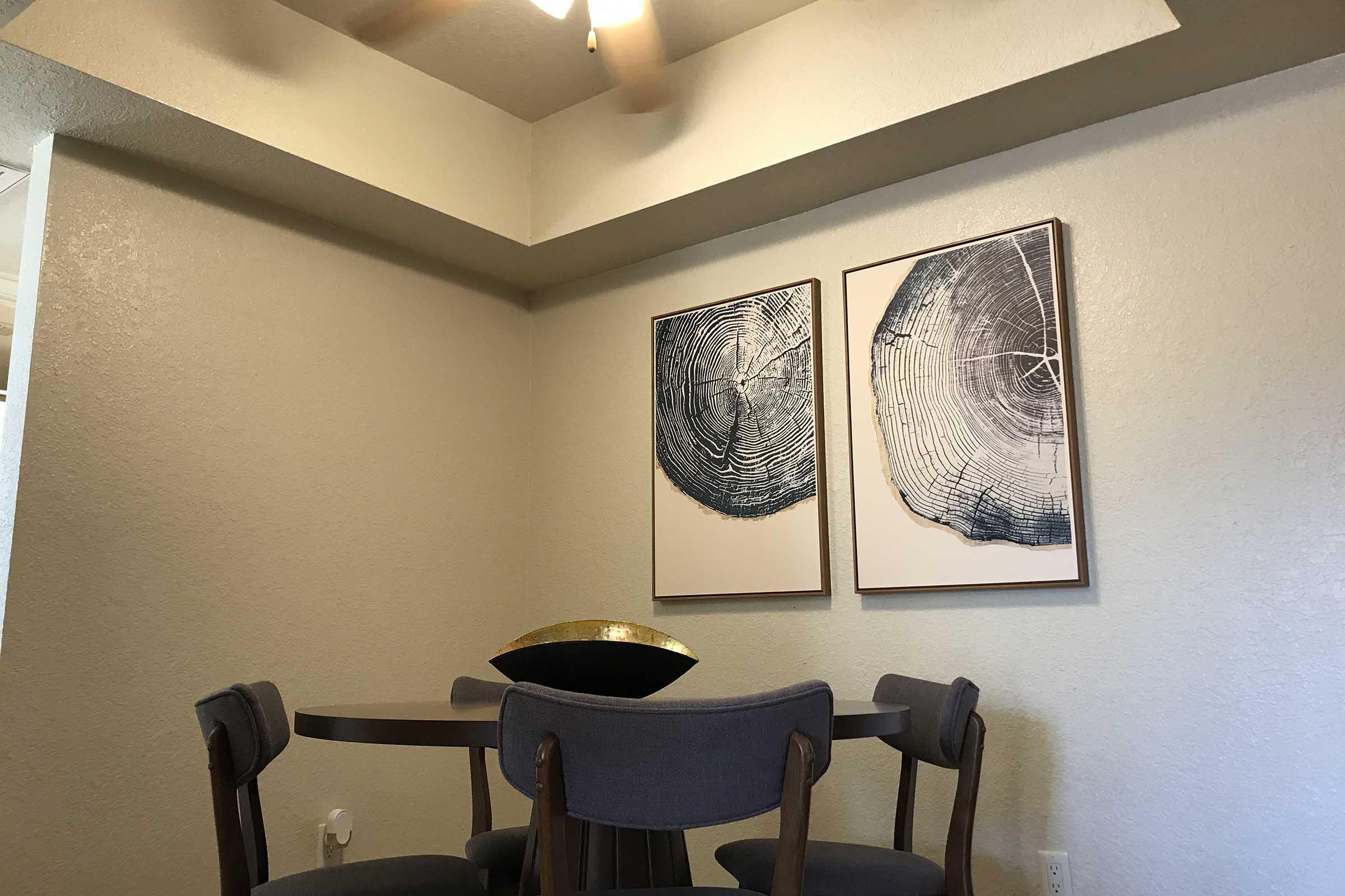 A cozy dining area featuring a round table with four gray chairs. Above the table, two framed art pieces depicting textured circular patterns are mounted on a light wall. A decorative bowl sits at the center of the table, while a ceiling fan is visible above. The ambiance is inviting and modern.