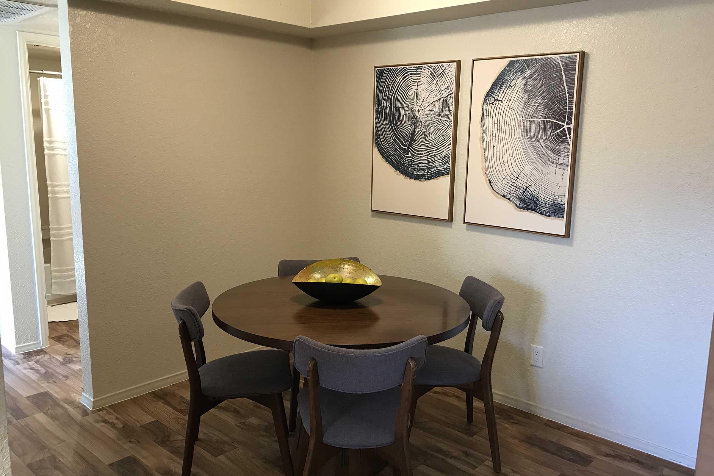 A modern dining area featuring a round wooden table surrounded by four upholstered chairs. Above the table are two framed abstract art pieces depicting circular patterns. A decorative bowl sits at the center of the table, and light-colored walls complement the wood flooring.