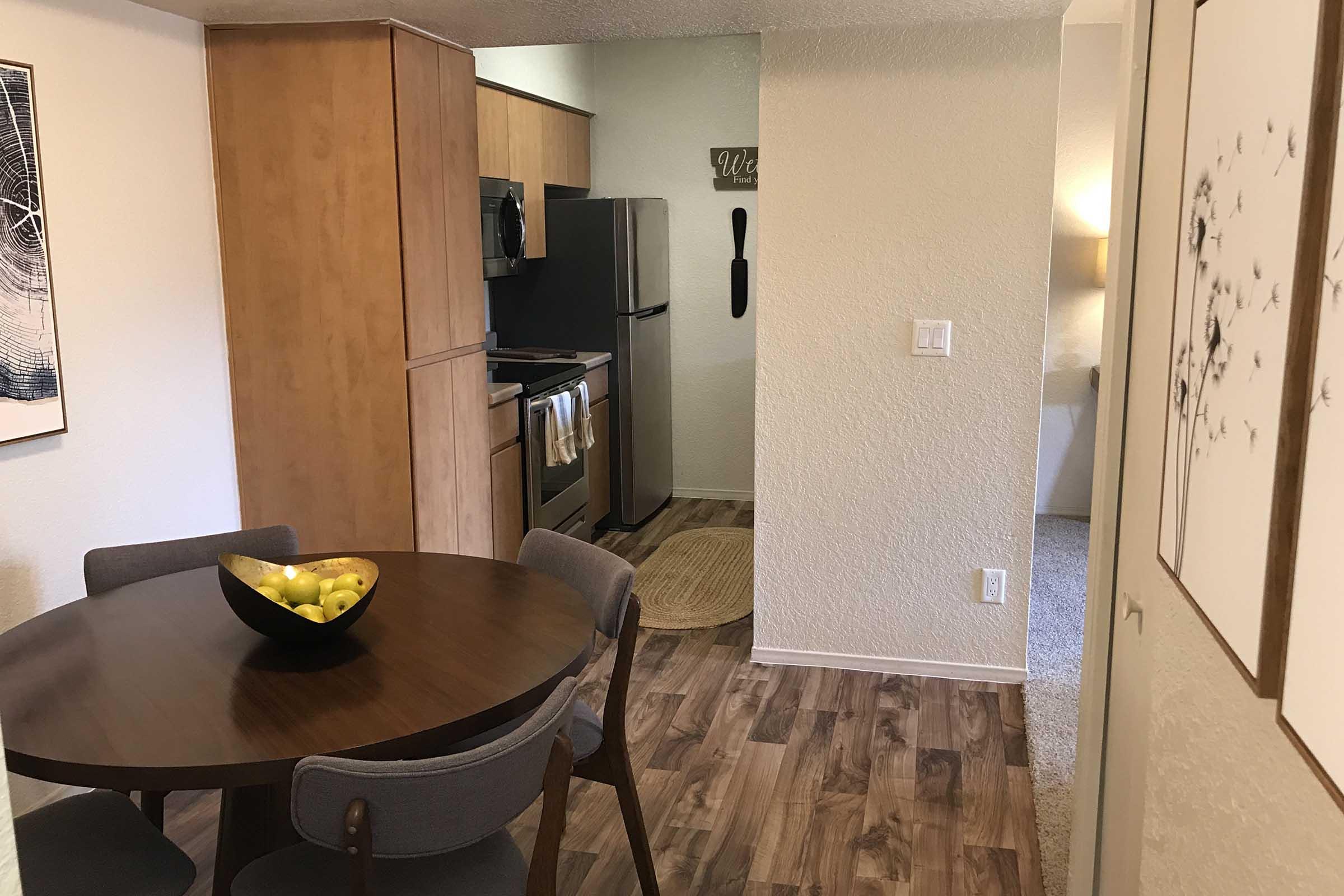 A modern kitchen area featuring light wood cabinetry, stainless steel appliances, and a round dining table with four chairs. A bowl of green apples sits at the center of the table. The space is well-lit and has neutral-colored walls and flooring, creating a cozy atmosphere.