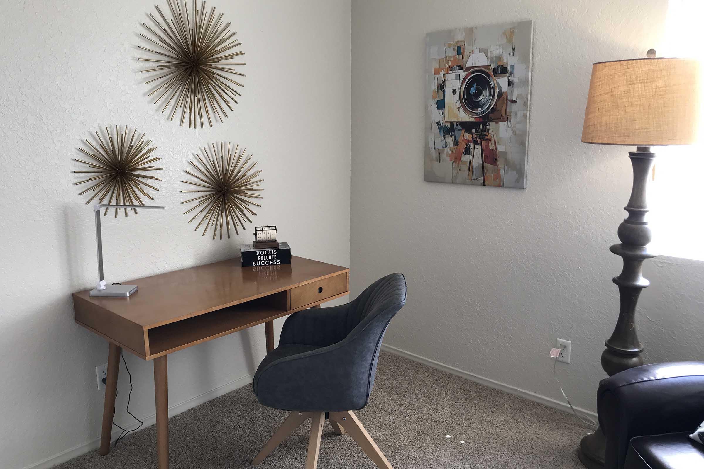 A cozy home office space featuring a wooden desk, a plush gray chair, and a decorative lamp beside a window. The walls display three sunburst wall decor pieces and a colorful abstract painting of a camera, adding a modern touch to the room's aesthetic.