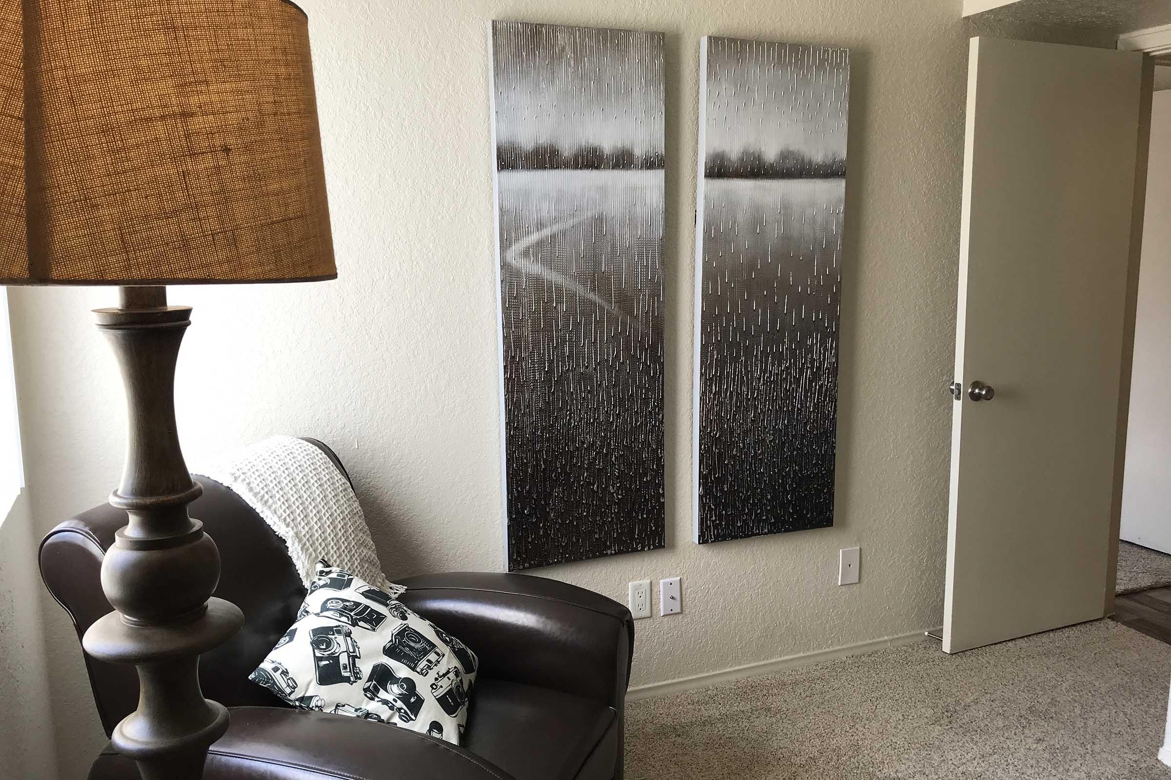 A corner of a room featuring a brown leather chair with a patterned pillow, a standing lamp with a textured shade, and two large abstract paintings depicting a tranquil, muted landscape on the wall. A closed door is visible in the background, adding to the cozy atmosphere.