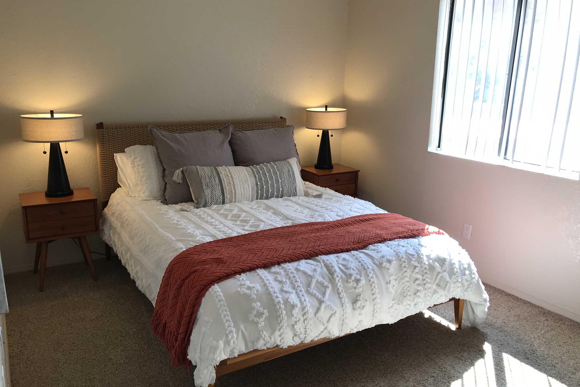 A cozy bedroom featuring a queen-sized bed with a white coverlet and decorative pillows, flanked by two bedside tables with black lamps. Natural light filters through large windows, illuminating the warm, neutral tones of the room. A soft area rug complements the overall inviting atmosphere.