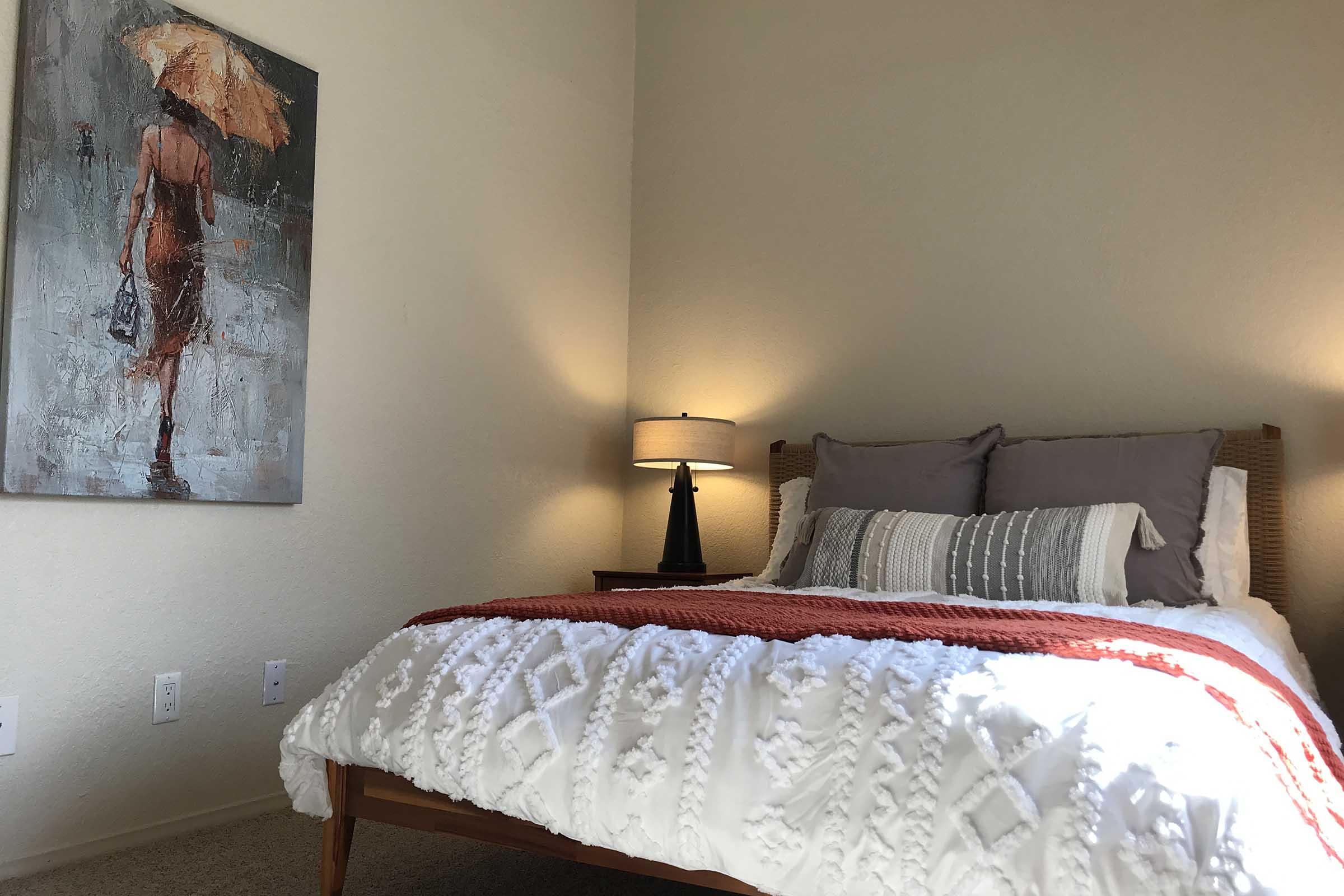 A cozy bedroom featuring a large bed with white and gray bedding, an orange throw blanket, and two decorative pillows. A bedside table with a lamp sits next to the bed. On the wall, there's an artistic painting of a figure holding an umbrella. The room has warm lighting and a neutral color palette.