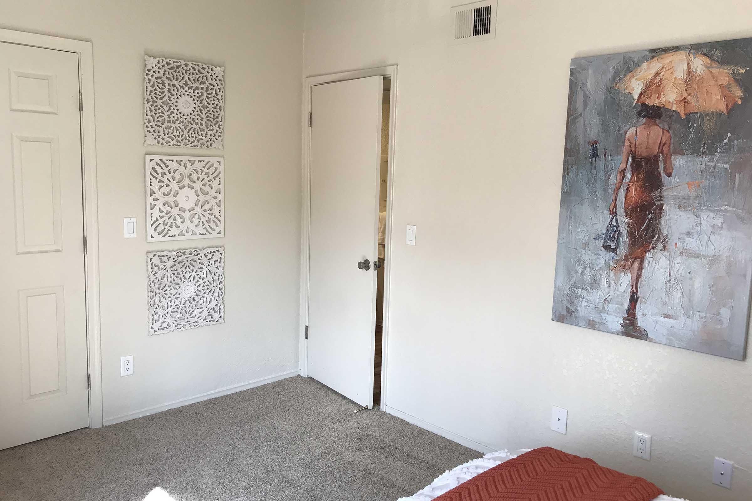 A cozy room featuring light-colored walls, a closed door, and decorative wall art. Three intricate wall panels are mounted on the left, while a large painting of a woman with an umbrella is on the right. The floor is carpeted, and a bed with a textured orange blanket is partially visible at the bottom.