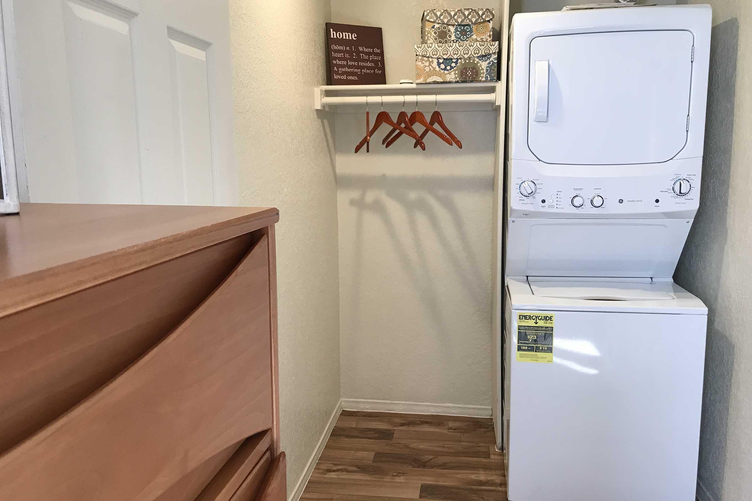 A small laundry area featuring a stacked washer and dryer, a shelf with decorative items, and a rod with three hangers. To the left, there's a wooden dresser with four drawers. The walls are light-colored, and the flooring resembles wood.