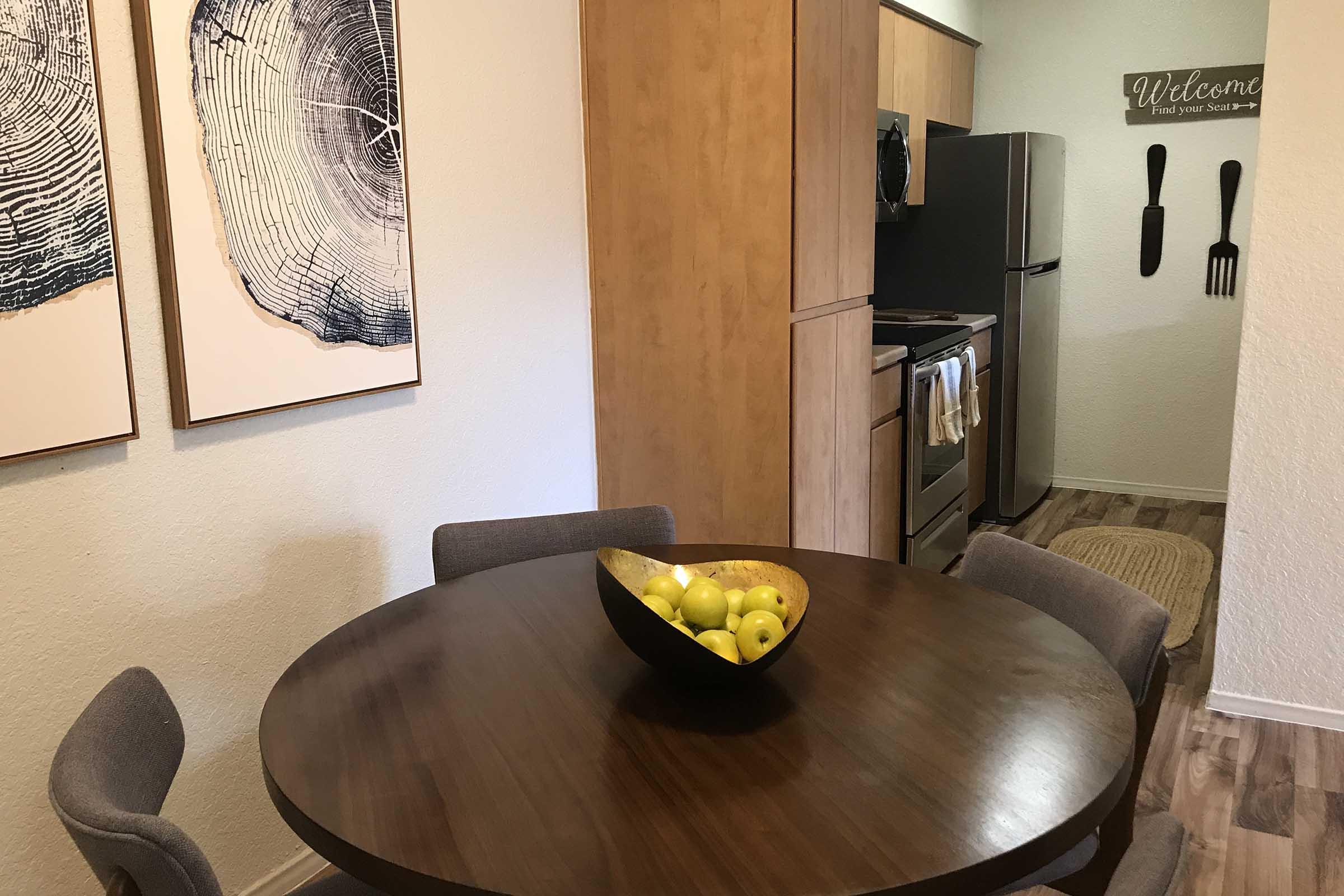 A cozy dining area featuring a round wooden table with a bowl of green apples at the center. Surrounding the table are four gray chairs. On the walls are two large framed artworks depicting tree rings. A kitchen area is visible in the background, with modern appliances and a welcoming sign.