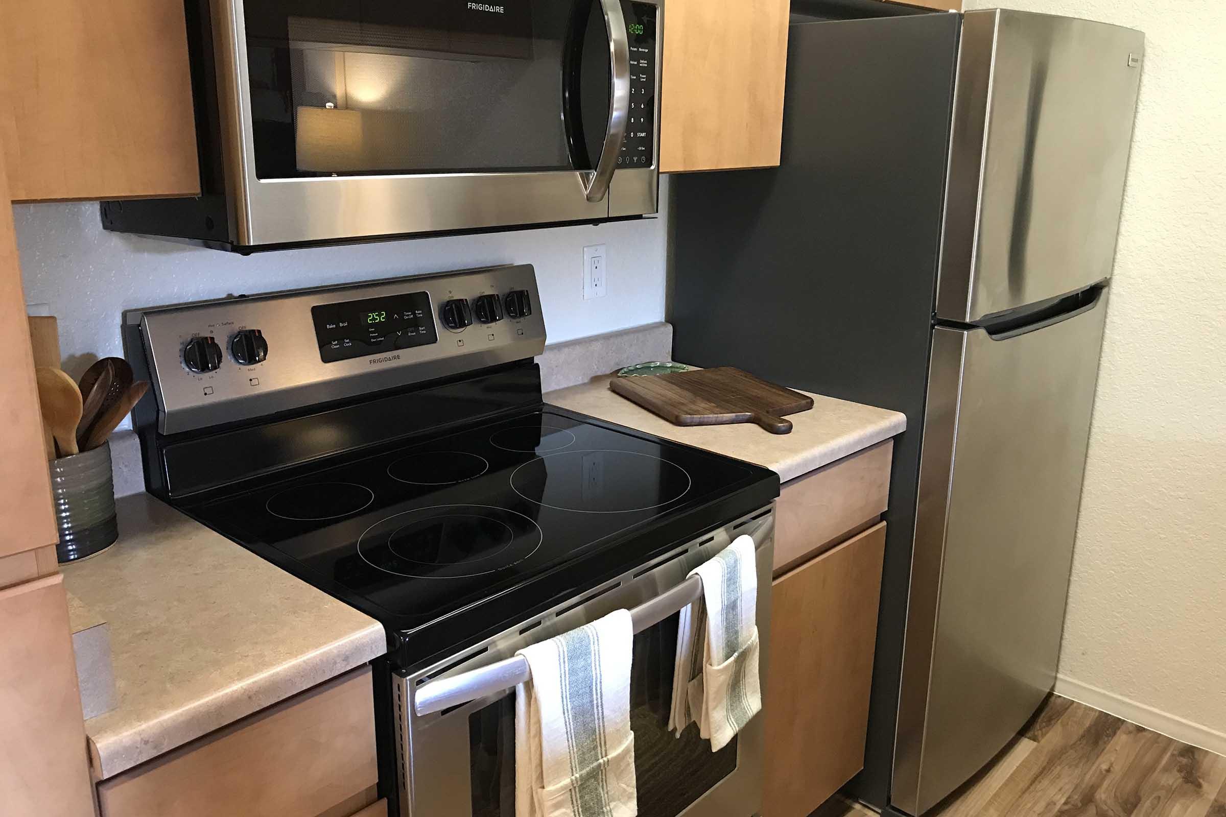 A modern kitchen featuring a stainless steel refrigerator, a built-in microwave above a black electric stove with a glass cooktop. There are wooden cabinets, a countertop with a cutting board, and kitchen utensils displayed next to the stove, along with a pair of hanging kitchen towels.