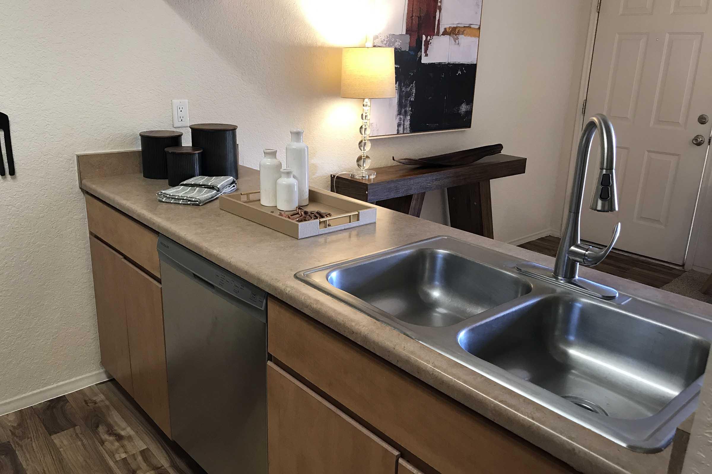 A modern kitchen countertop with a double sink and a dishwasher. Decorative items include a tray with bottles and a small bowl of nuts, alongside a stylish lamp providing warm lighting. The background shows a simple wooden table and a door leading outside.