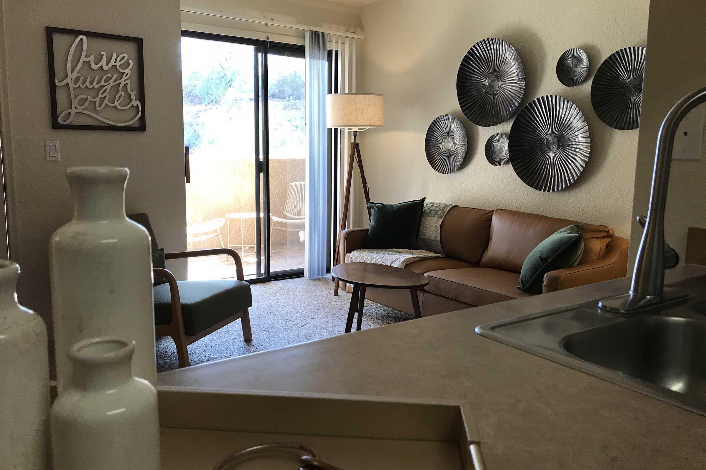 A cozy living room featuring a brown sofa with green and gray pillows, a round wooden coffee table, and a decorative wall with metal circular art pieces. There is a view of a balcony through sliding glass doors, and a lamp stands beside a chair. Decorative vases are placed on a counter in the foreground.