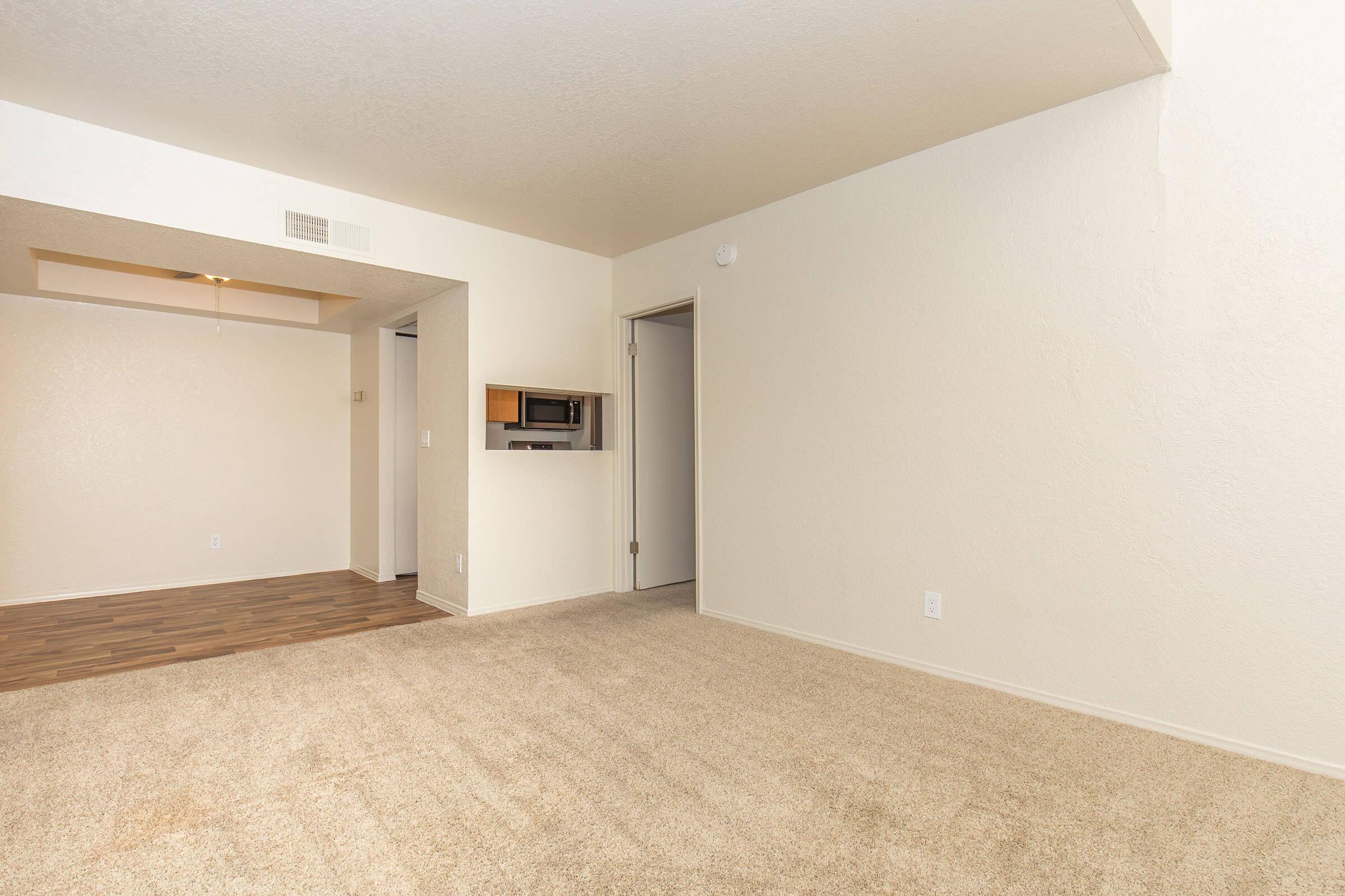 A spacious, well-lit living area with beige walls and carpet, featuring a doorway leading to another room. On the left, there's a small opening with a window, possibly a pass-through to a kitchenette. The area is minimalistic, creating an inviting and open atmosphere.