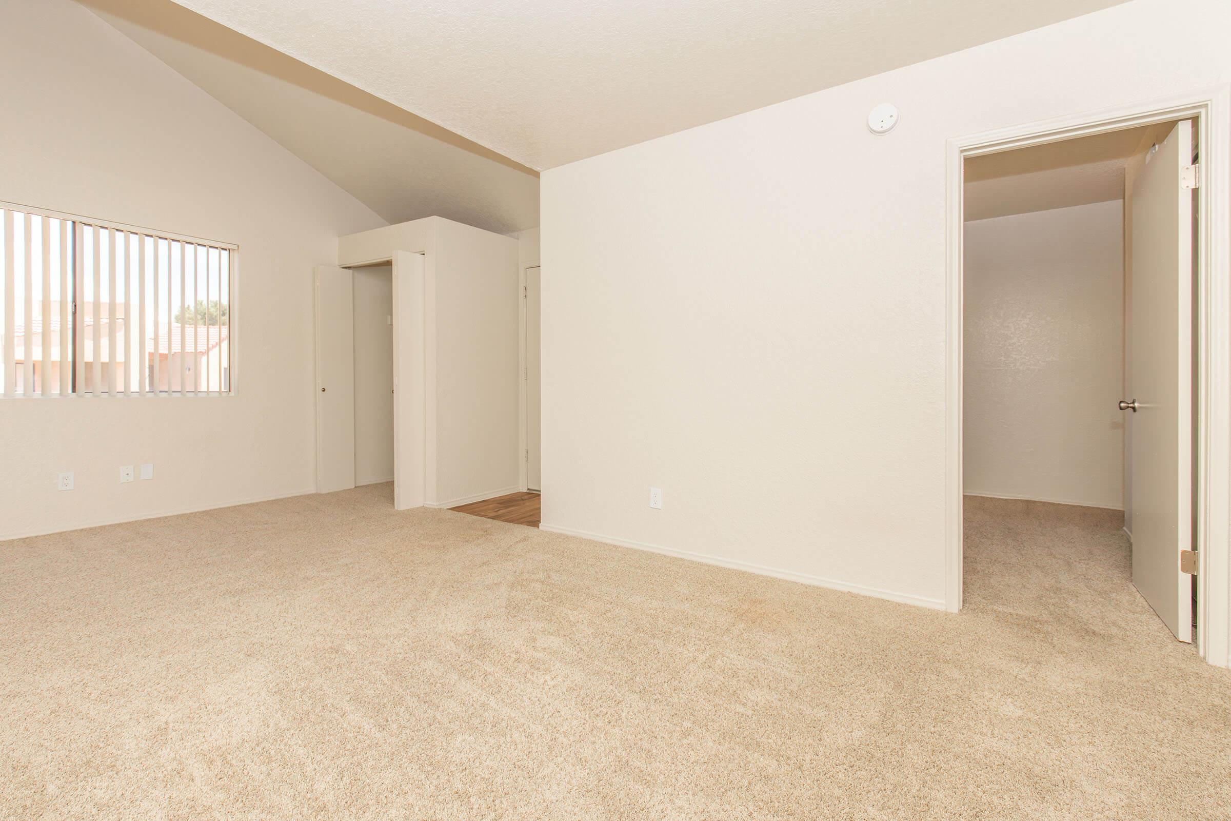 A spacious, empty room with light beige carpet and white walls. The room features a high ceiling, a large window with vertical blinds, and an open doorway leading to another room. There are no furnishings, creating a bright and airy atmosphere.