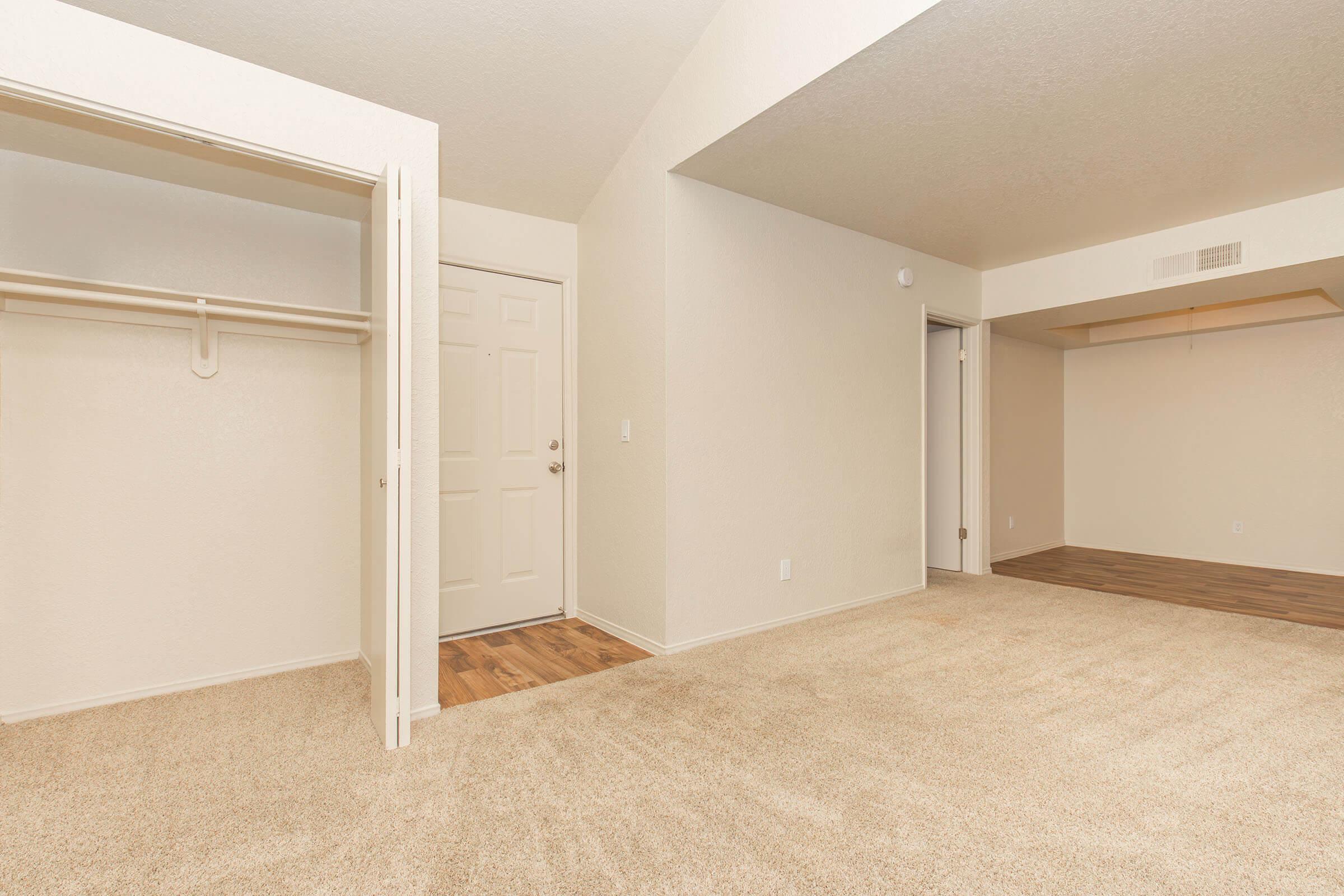 A well-lit, empty room with beige walls and carpeted flooring. There is a doorway leading to another room, a closet with sliding doors on the left, and a corner without any furniture. The space appears spacious and ready for decoration.