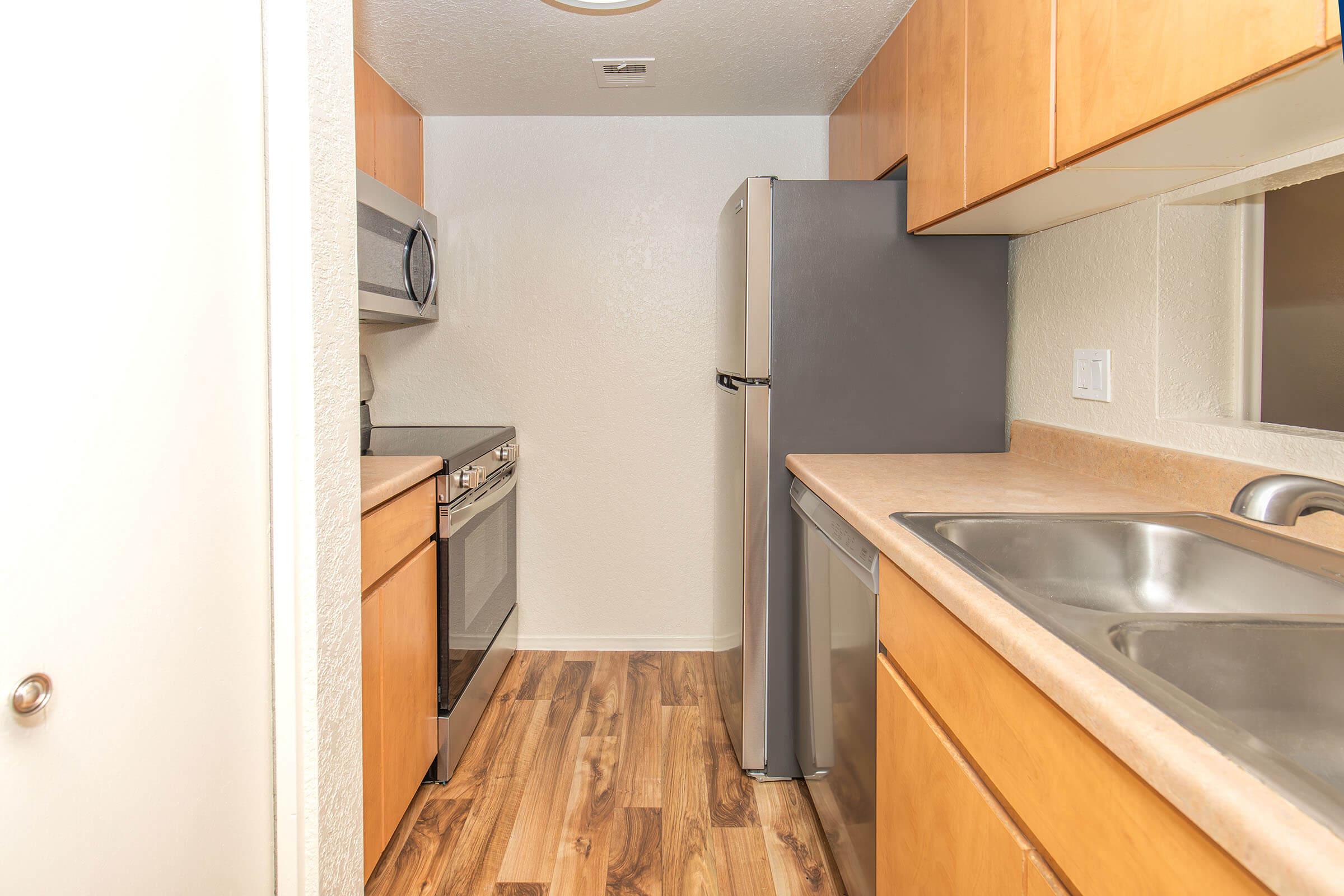 A modern kitchen featuring wooden cabinetry, stainless steel appliances including a microwave, oven, and refrigerator, a dual sink, and laminate flooring. The space is well-lit with a neutral color scheme and an open layout.