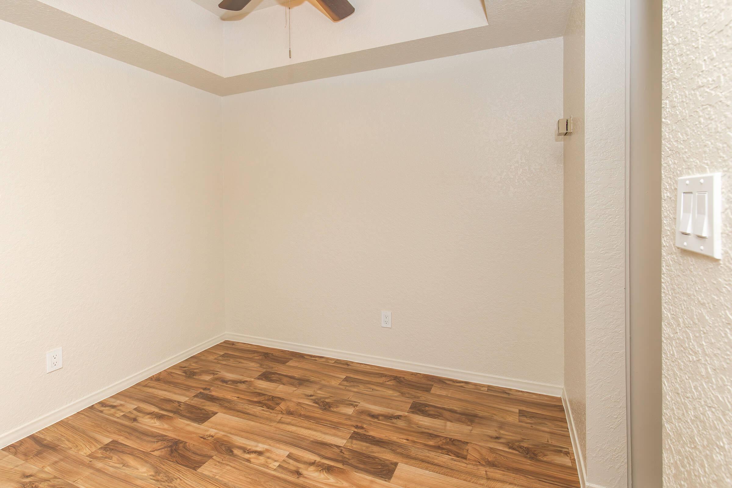 Empty interior of a room with light-colored walls and wooden flooring. A ceiling fan is visible, and there is a light switch on the right side. The space looks well-lit and clean, ready for furniture or decoration.