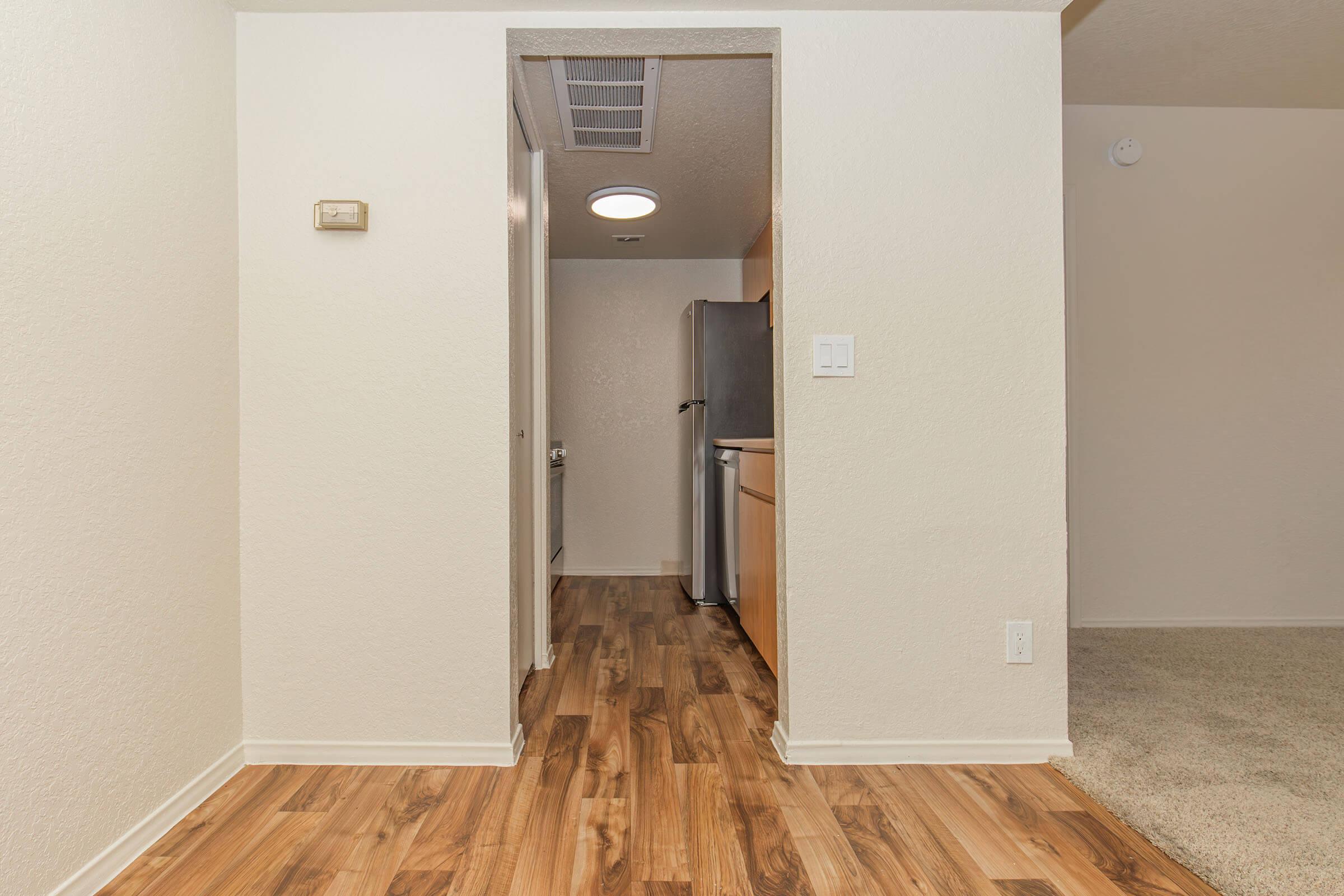 Interior view of a hallway leading to a kitchen area, featuring light-colored walls, wooden flooring, and an open layout. To the left, a doorway leads to the kitchen, which contains stainless steel appliances. A small hallway light and vent are visible on the ceiling, and a carpeted area is seen on the right.