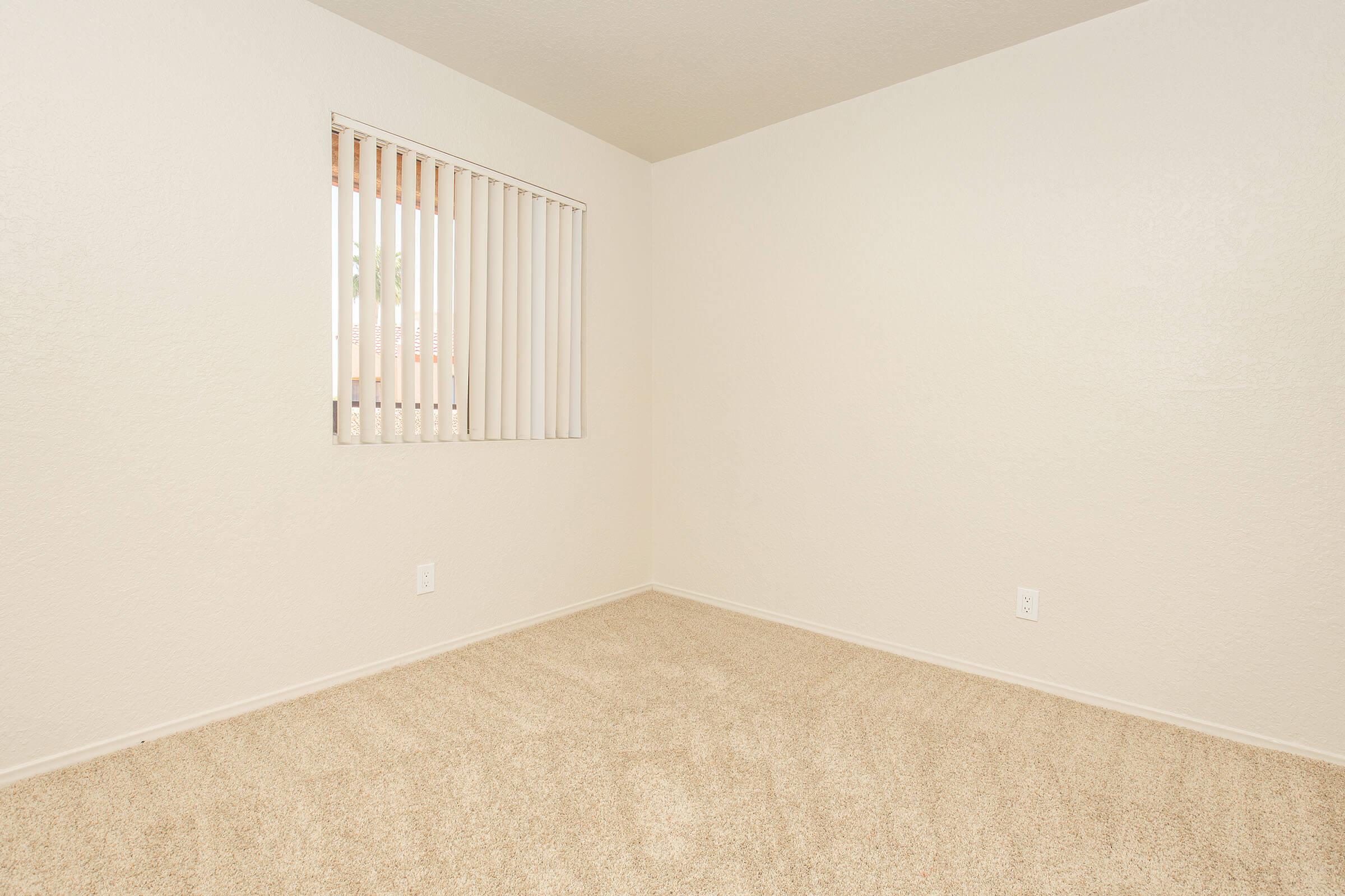 An empty room with beige carpet, white walls, and a window featuring vertical blinds. The room appears well-lit and spacious, with no furniture or decorations present.