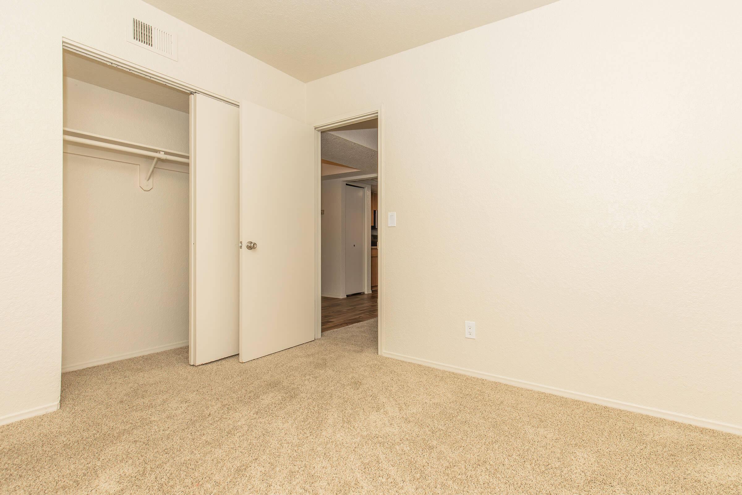 A clean, empty room featuring beige carpet, a closed closet with a sliding door, and two open doors leading to adjacent spaces. The walls are painted in a light color, contributing to a bright, spacious feel. The overall atmosphere is neutral and inviting.