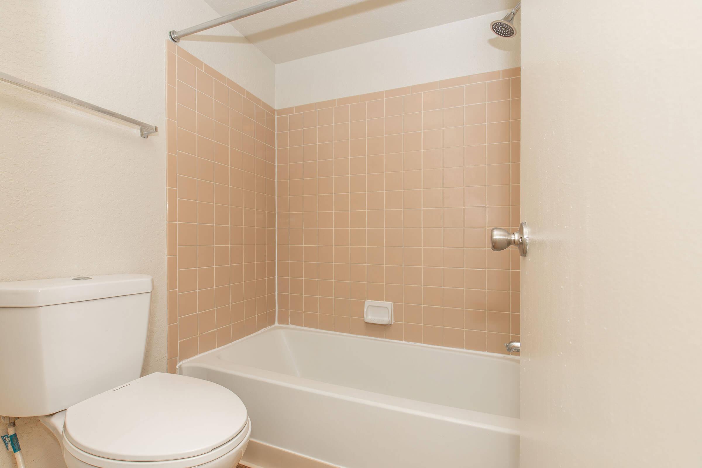 A clean bathroom featuring a bathtub with a pink tiled surround, a toilet, and a showerhead. The walls are painted white, adding to the bright appearance of the space. The bathroom design emphasizes simplicity and functionality.