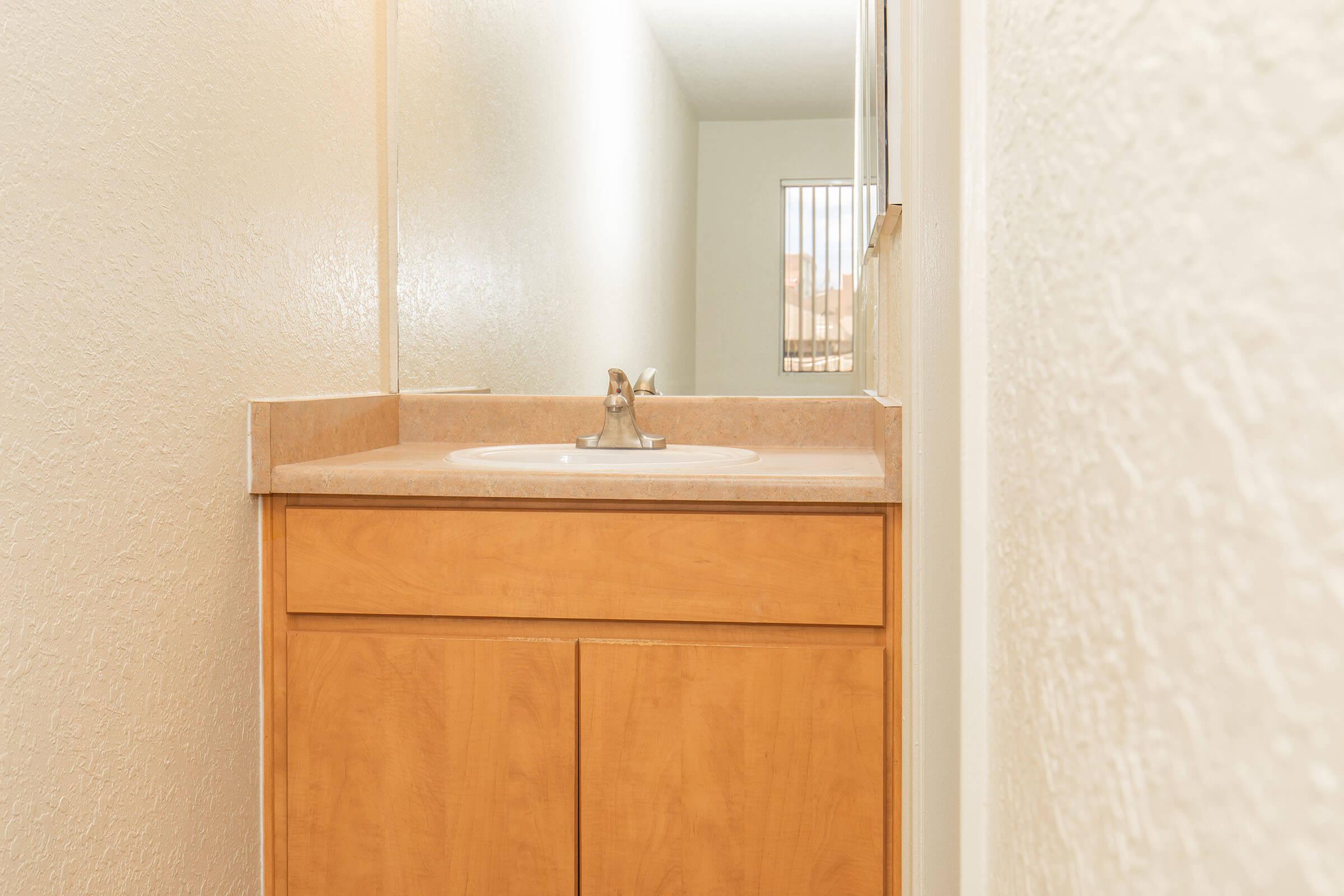 A small bathroom featuring a wooden cabinet under a sink with a faucet. The walls are painted in a light color, and there is a mirror above the sink. Natural light is coming from a window, and the space appears tidy and well-maintained.