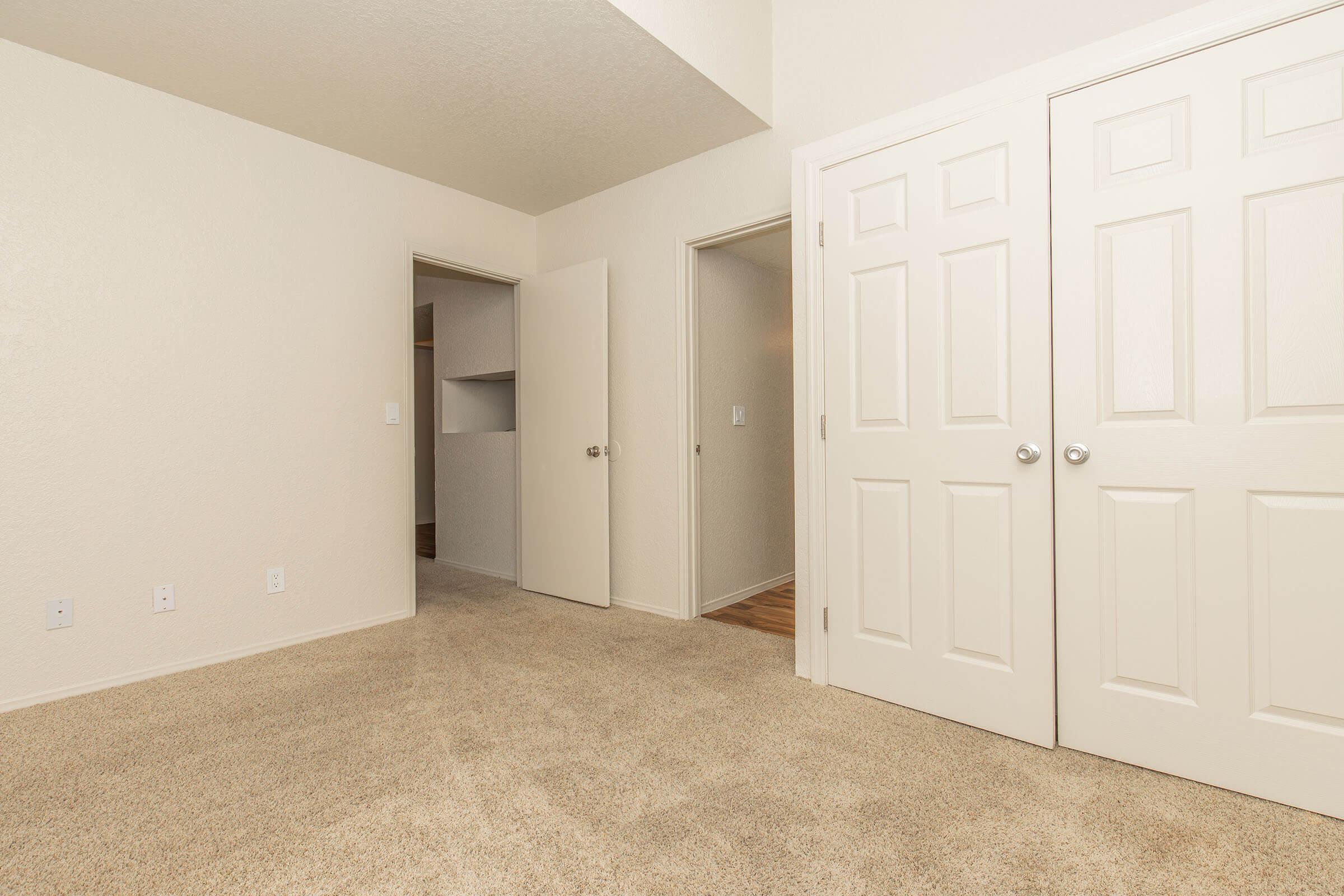 A well-lit, minimalistic room with beige carpet and light-colored walls. Two closed doors are visible on the right, and an open door leads to another room. The space feels clean and empty, ideal for various uses.