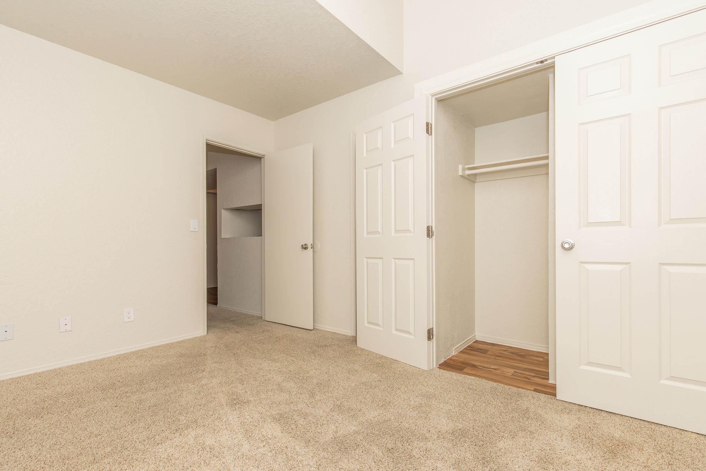 A spacious, lightly colored bedroom featuring beige carpet. Two doors lead out, while a third door opens to a closet with a shelf. The walls are painted a soft hue, creating an inviting atmosphere. There's ample natural light, making the room feel bright and airy.