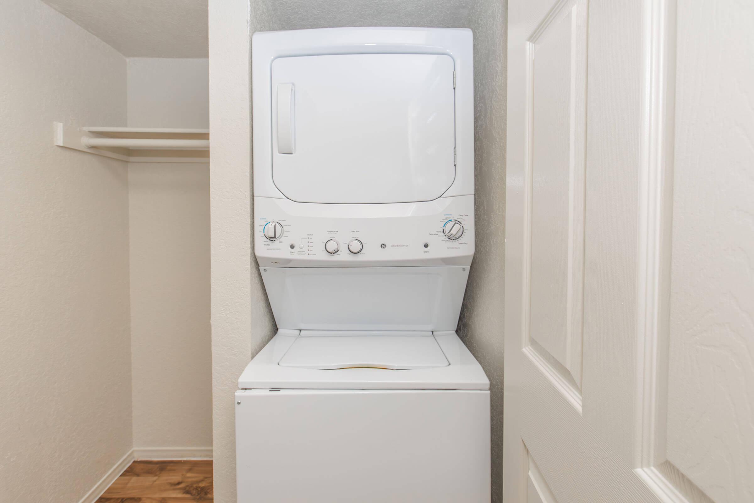 A stacked washer and dryer set positioned in a small laundry space, with a light-colored wall and a closet rod visible in the background. The floor features a wooden design, and the laundry appliances have control dials and buttons on the front.