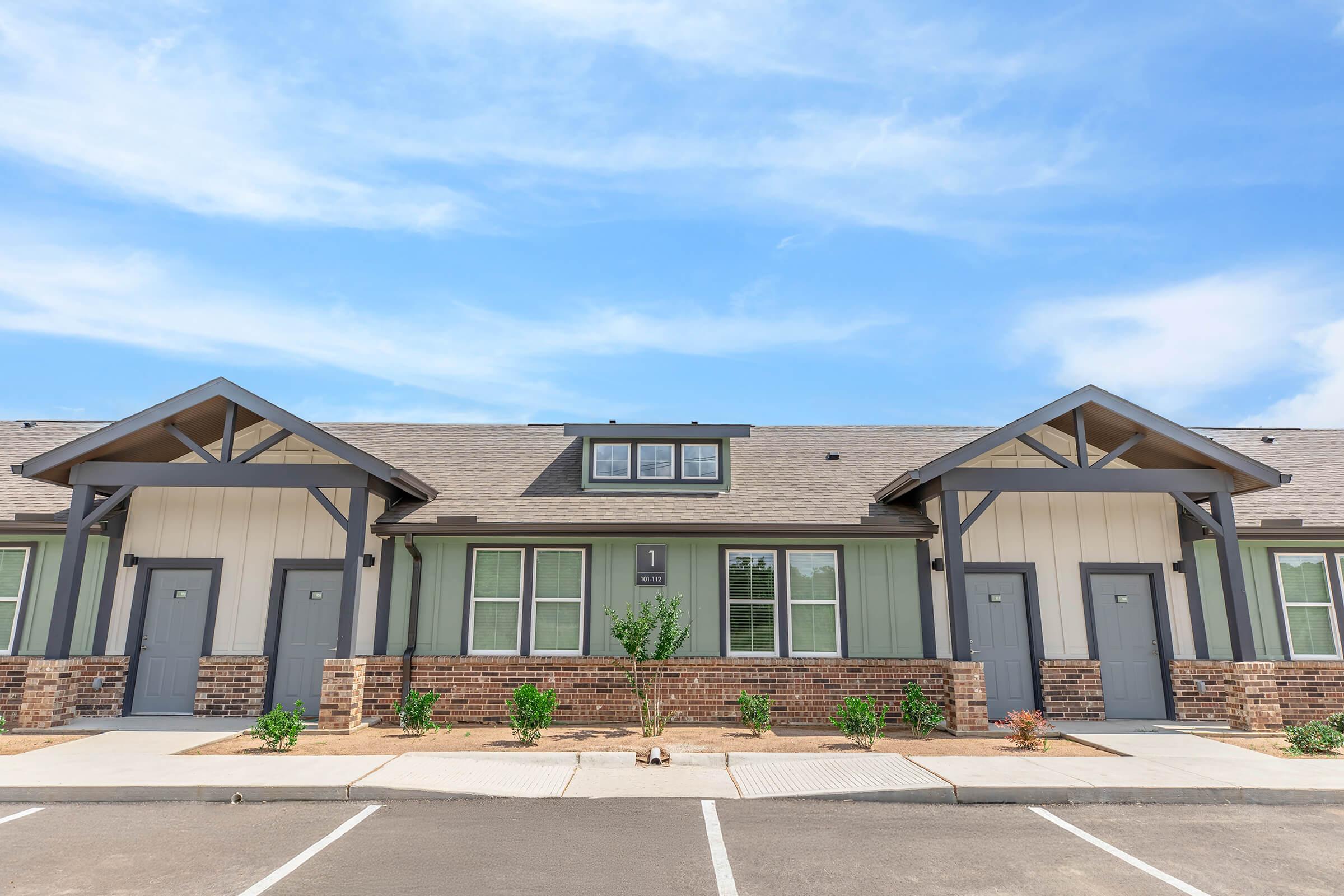 A row of modern green and beige townhomes with a brick facade, featuring gray doors and stylish rooflines. The scene is set under a clear blue sky, with small shrubs and landscaping in front of each unit.