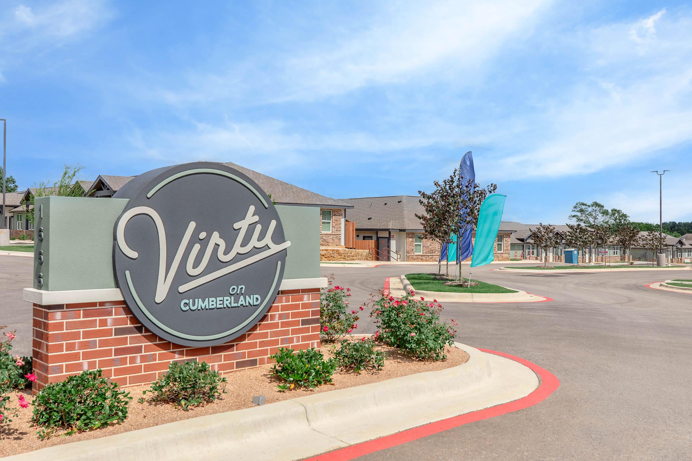 Sign for "Virtu on Cumberland" displayed prominently at the entrance of a residential community. The area features landscaped gardens with flowers and trees, modern architecture in the background, and a clear blue sky. The sign includes a logo and is set on a brick base.