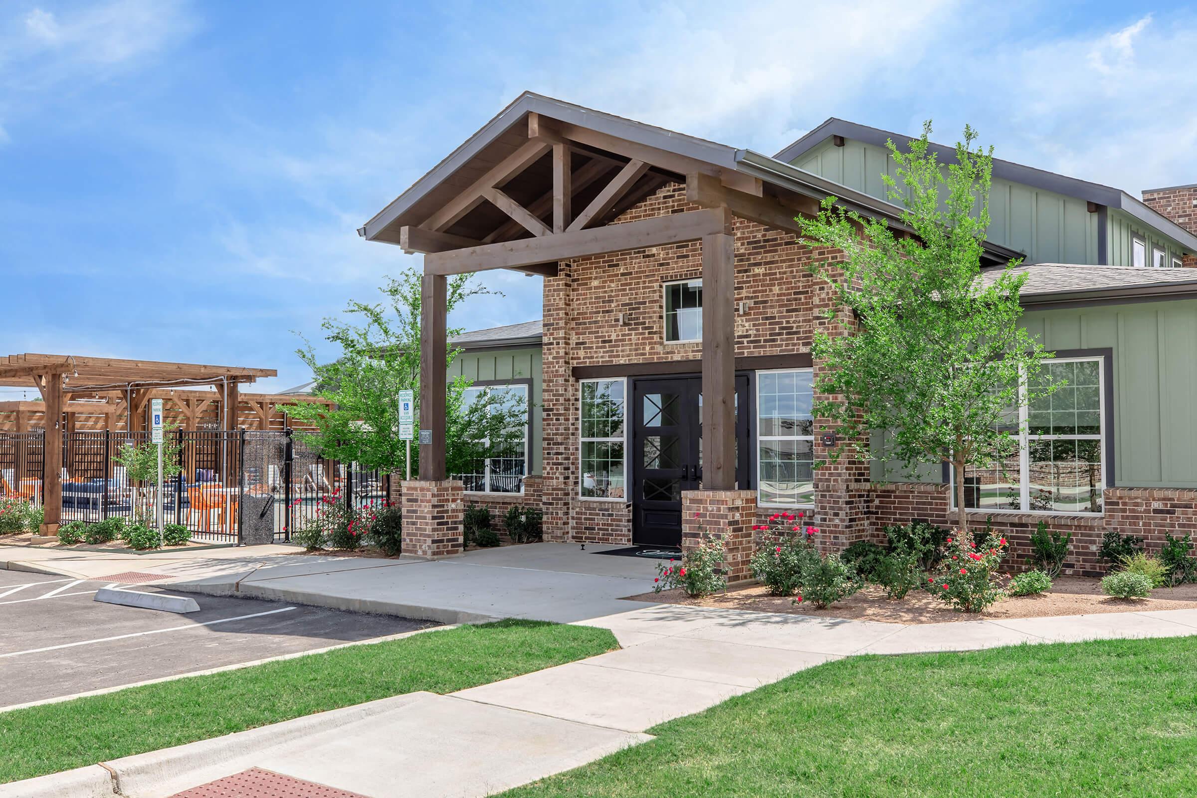A modern brick building with a large covered entrance, surrounded by greenery and neatly landscaped flower beds. A parking area is visible in front, and a wooden structure can be seen in the background, suggesting a recreational area. The sky is bright and clear.